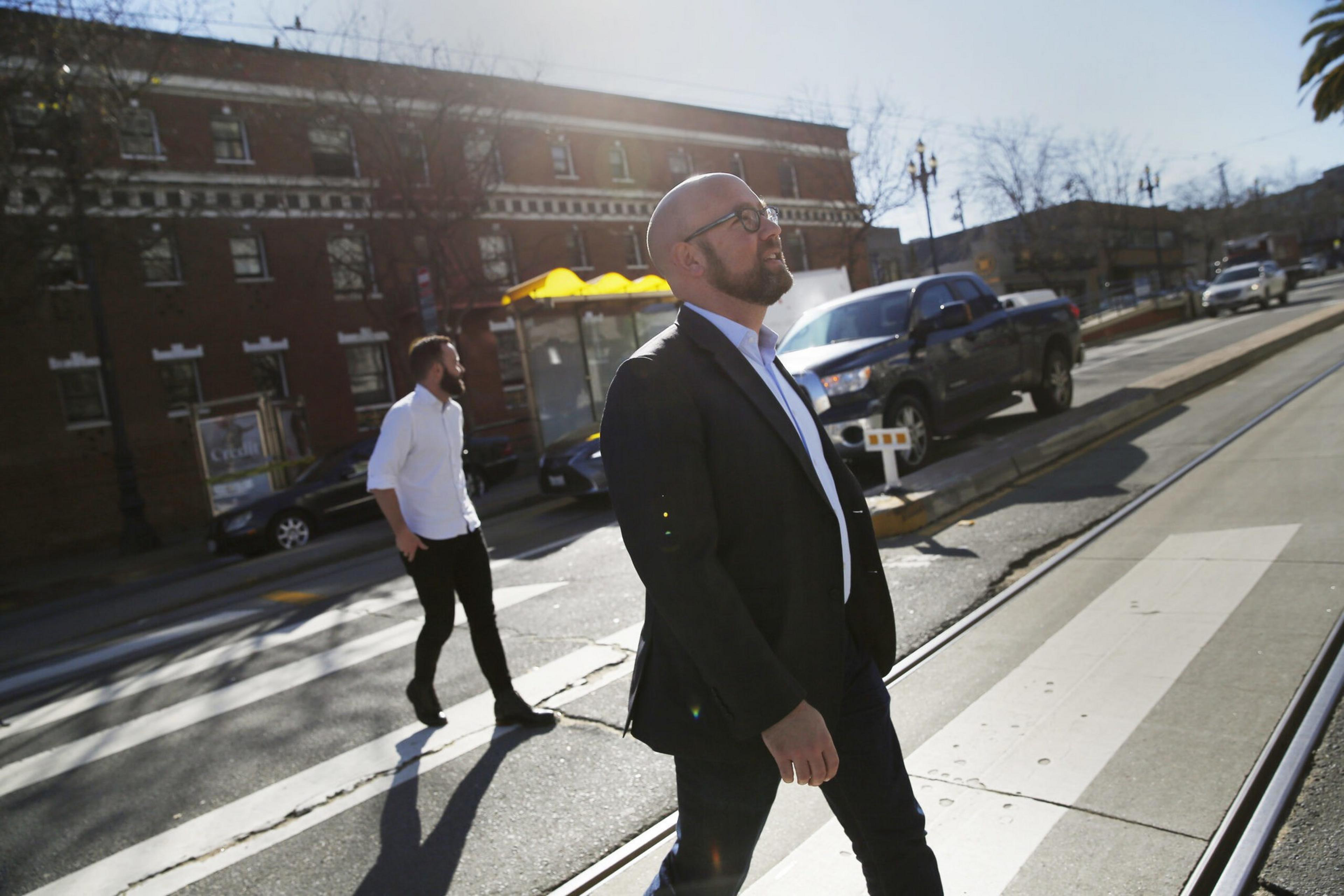 A man in a black suit and glasses walks on a crosswalk while another man in a white shirt walks behind him on a sunny urban street.