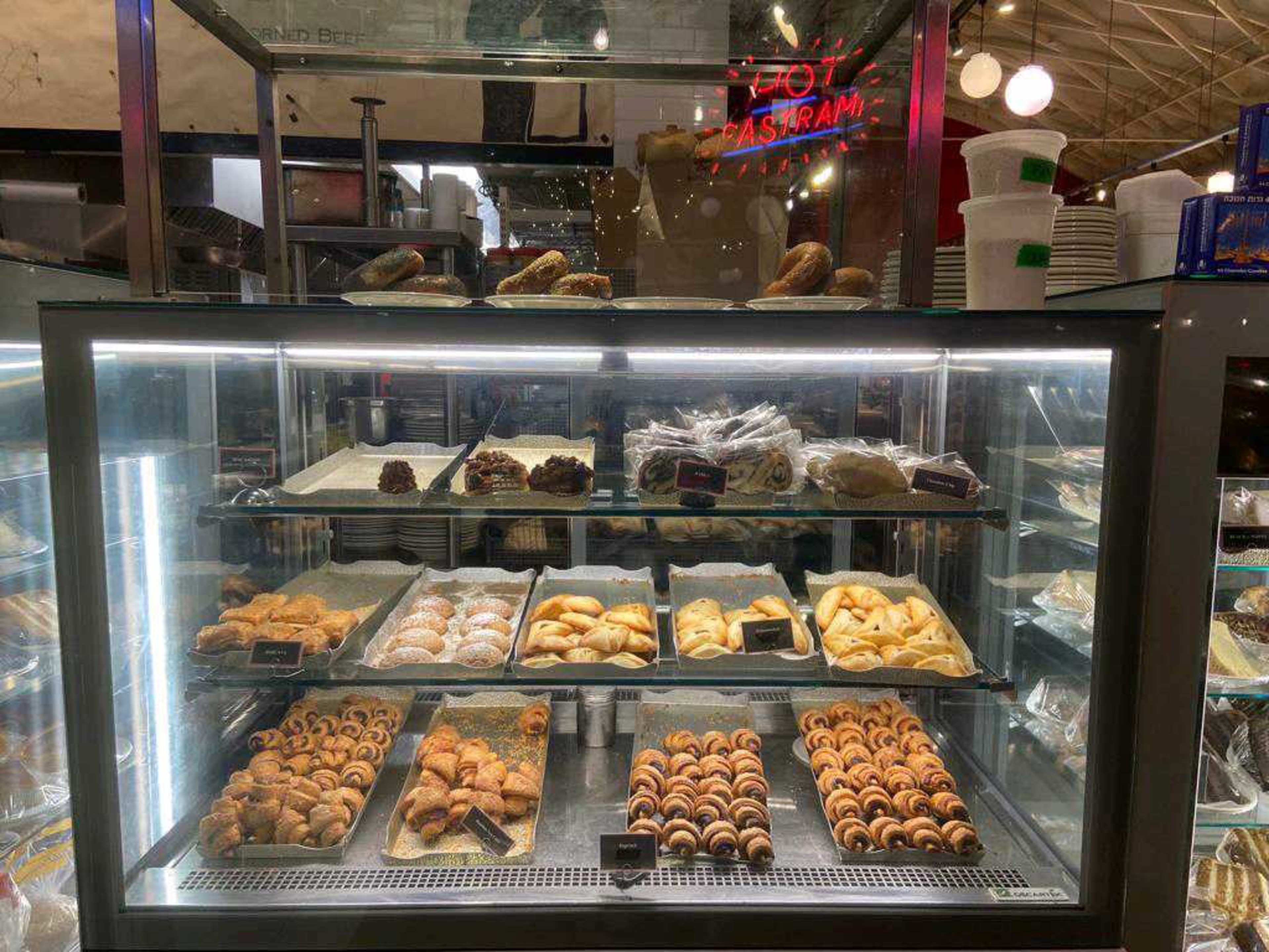 A bakery display case contains bagels on top, various pastries including rugelach, cookies, croissants, and rugelach rolls on three illuminated shelves.