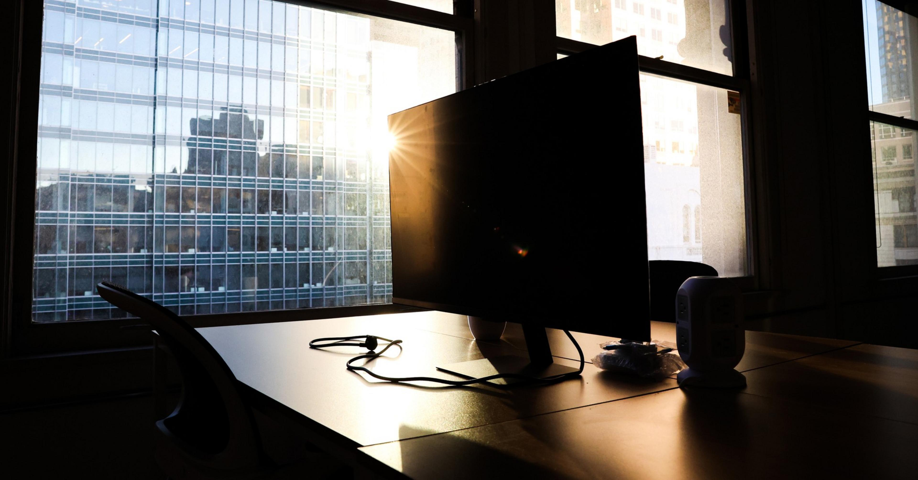 A computer monitor sits on a desk with cables, backlit by sunlight streaming through large windows showing city buildings.