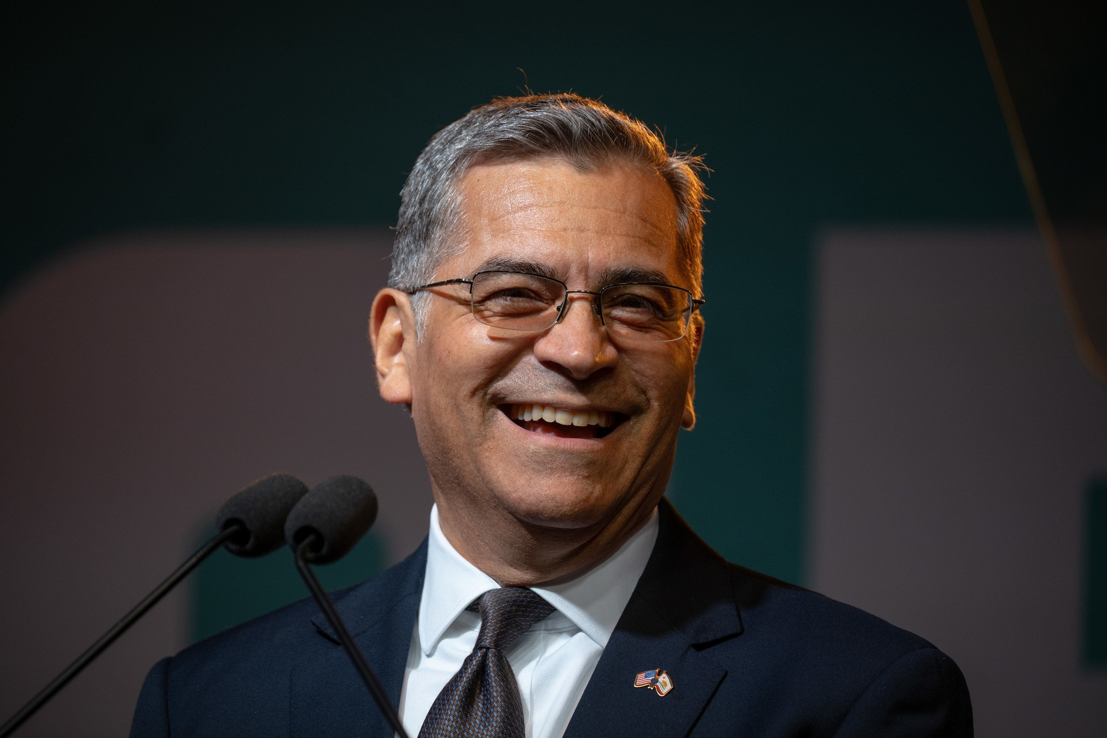 A man with gray hair and glasses smiles warmly while wearing a dark suit, white shirt, and tie, standing near two microphones.