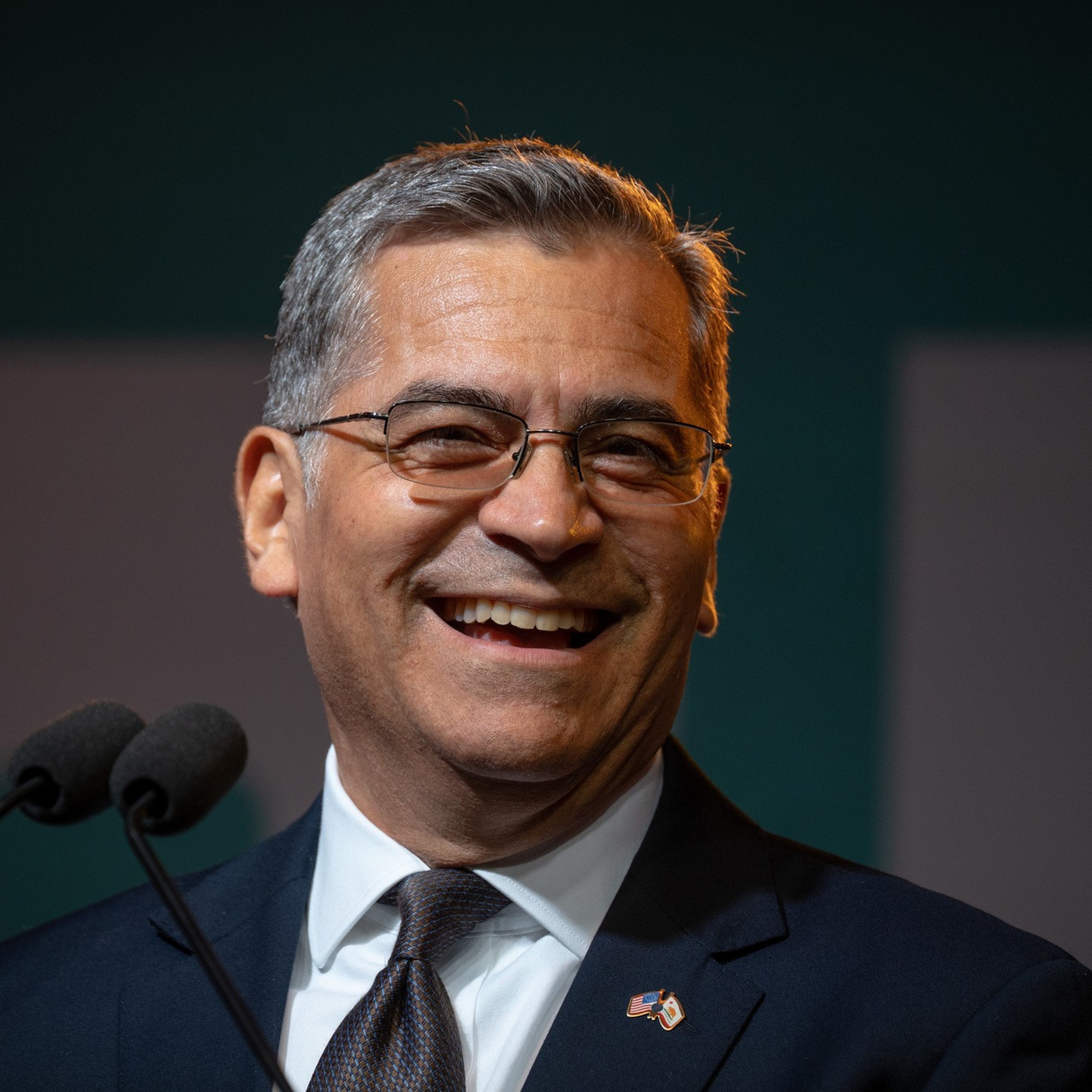 A man with gray hair and glasses smiles warmly while wearing a dark suit, white shirt, and tie, standing near two microphones.