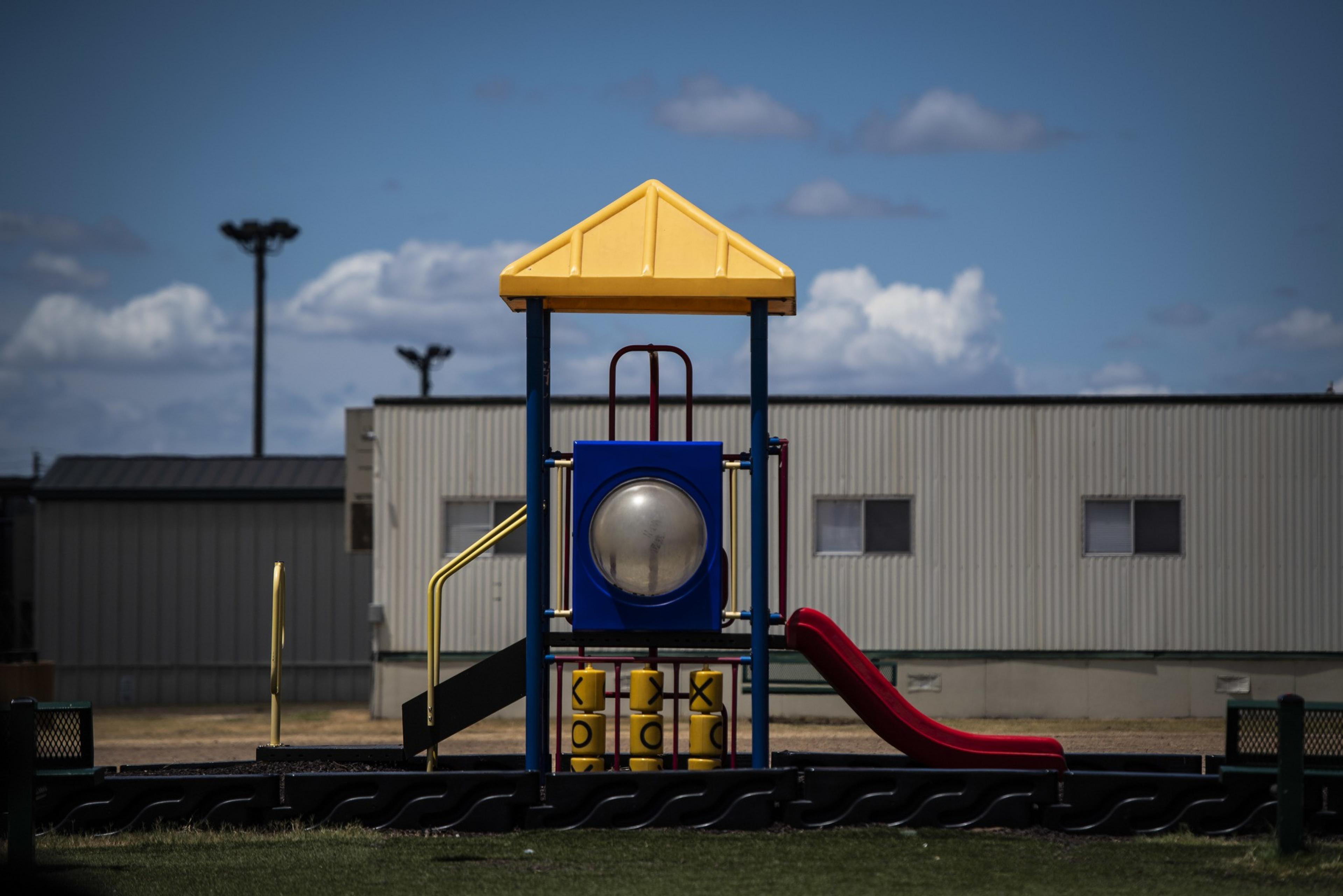 A playground structure with a yellow roof, blue panel, a spherical window, tic-tac-toe cylinders, a yellow ladder, and a red slide, against a cloudy sky and a building.