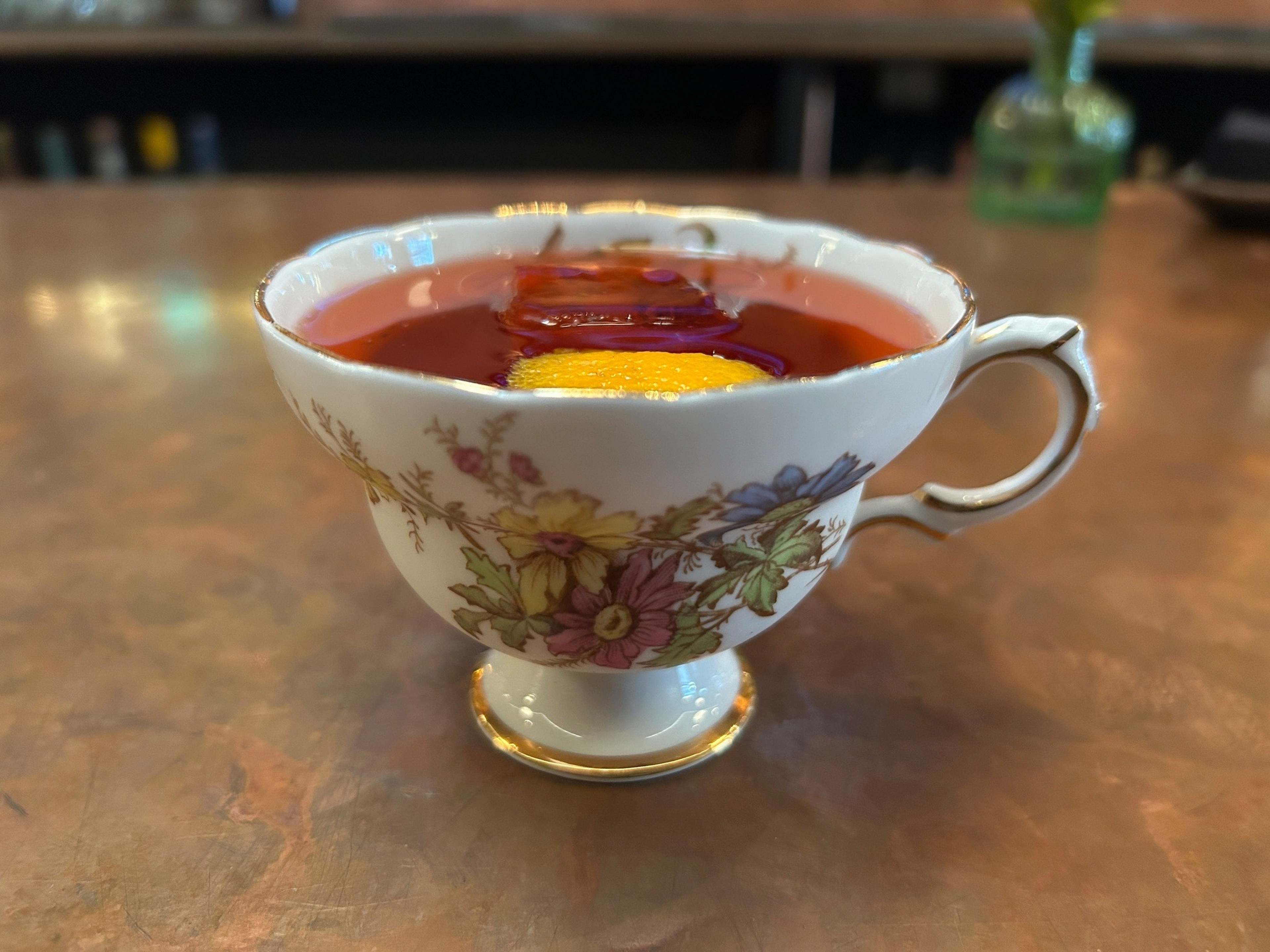 A porcelain teacup with floral designs holds red tea with a slice of lemon floating on top, set on a polished wooden surface.