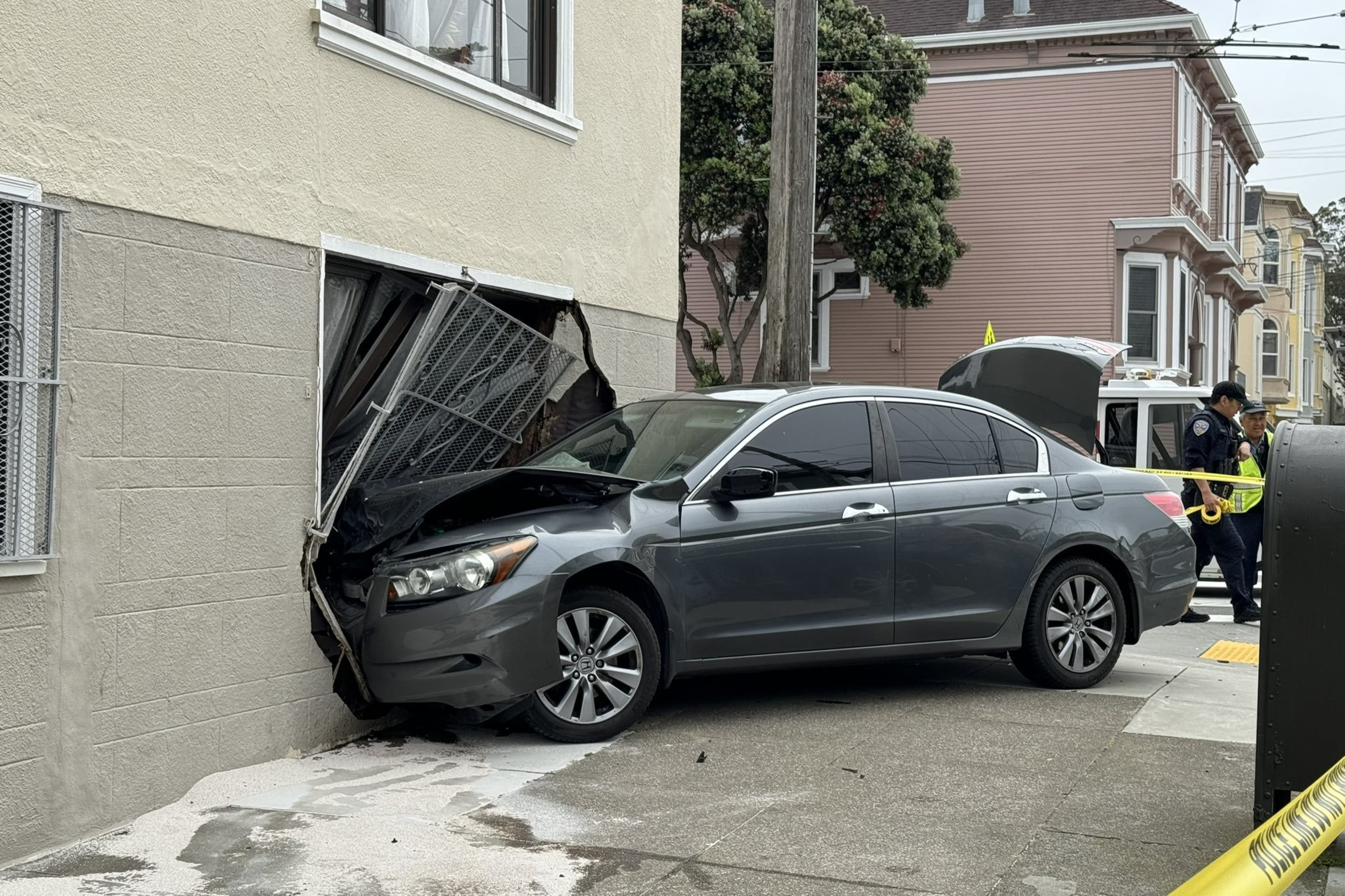 A gray car has crashed into the side of a building, damaging a garage door. A police officer is at the scene, inspecting the damage.
