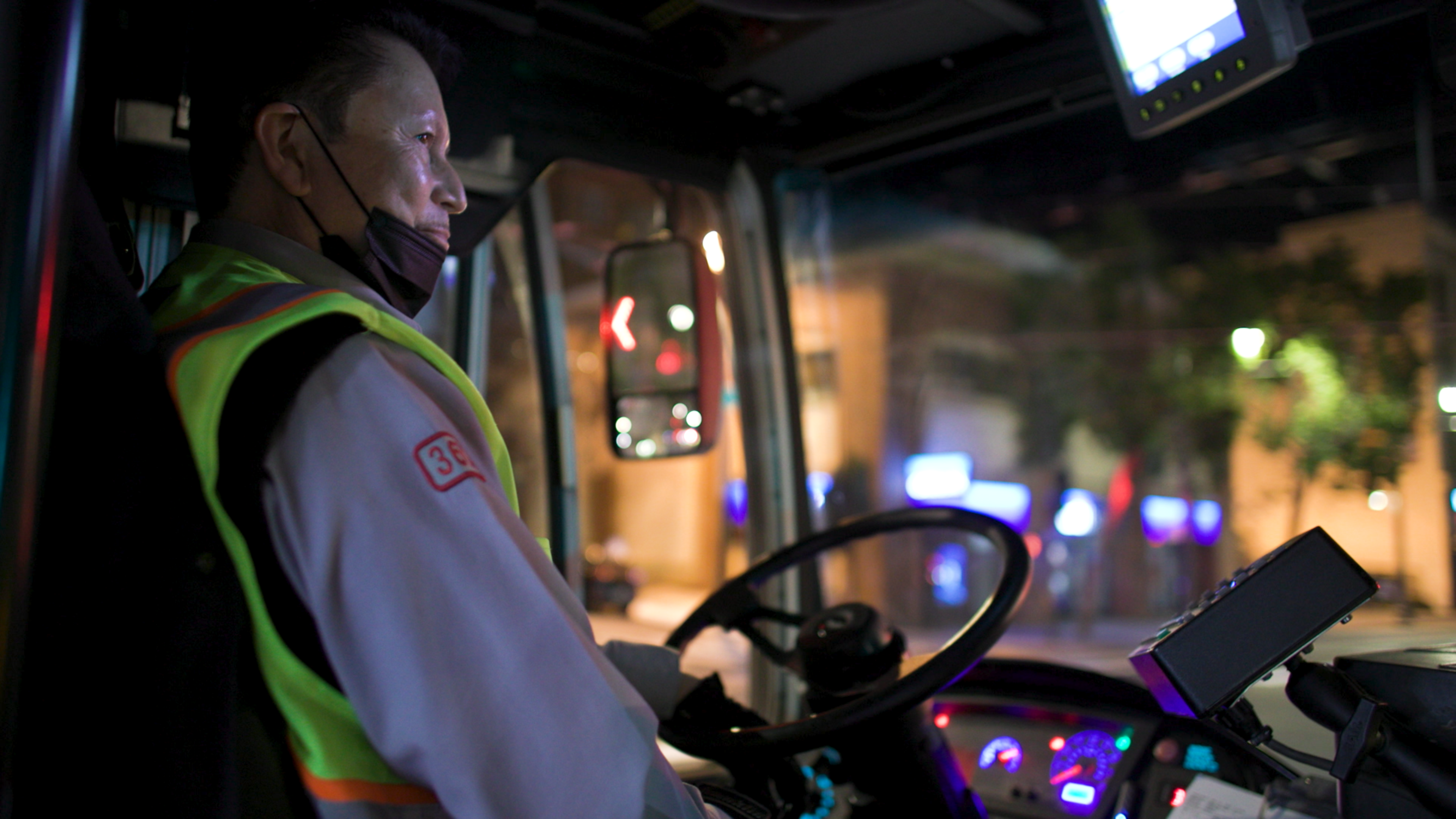 A bus driver wearing a reflective vest and face mask sits at the wheel of a bus at night, with illuminated dashboard and streetlights outside.