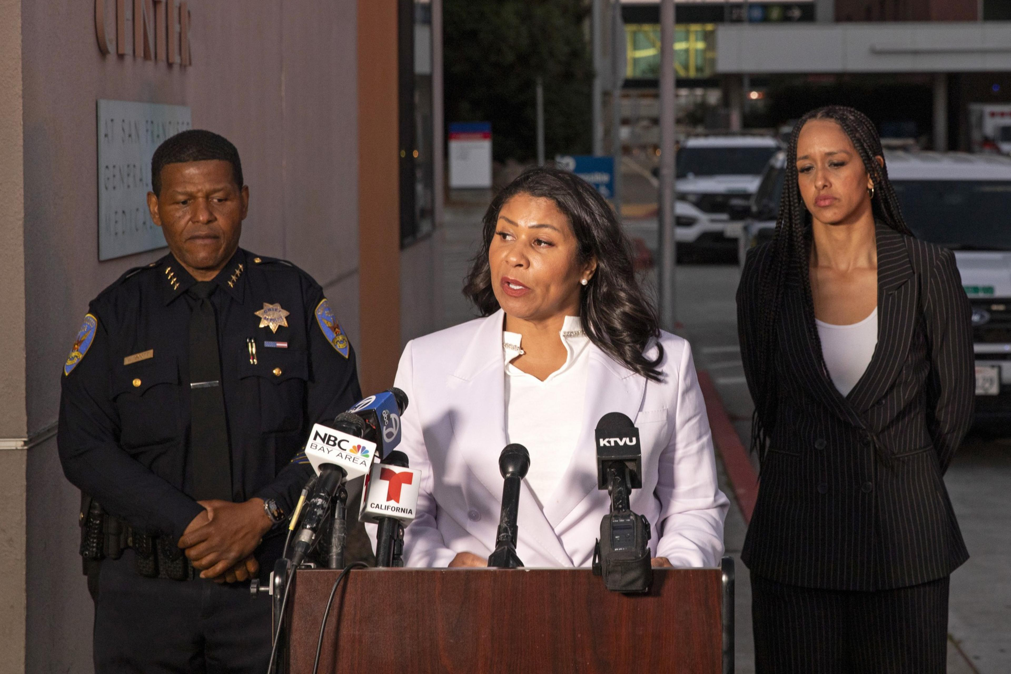 Three individuals, including a uniformed officer, stand behind a podium with multiple microphones set up for a press conference held outside near a building entrance.