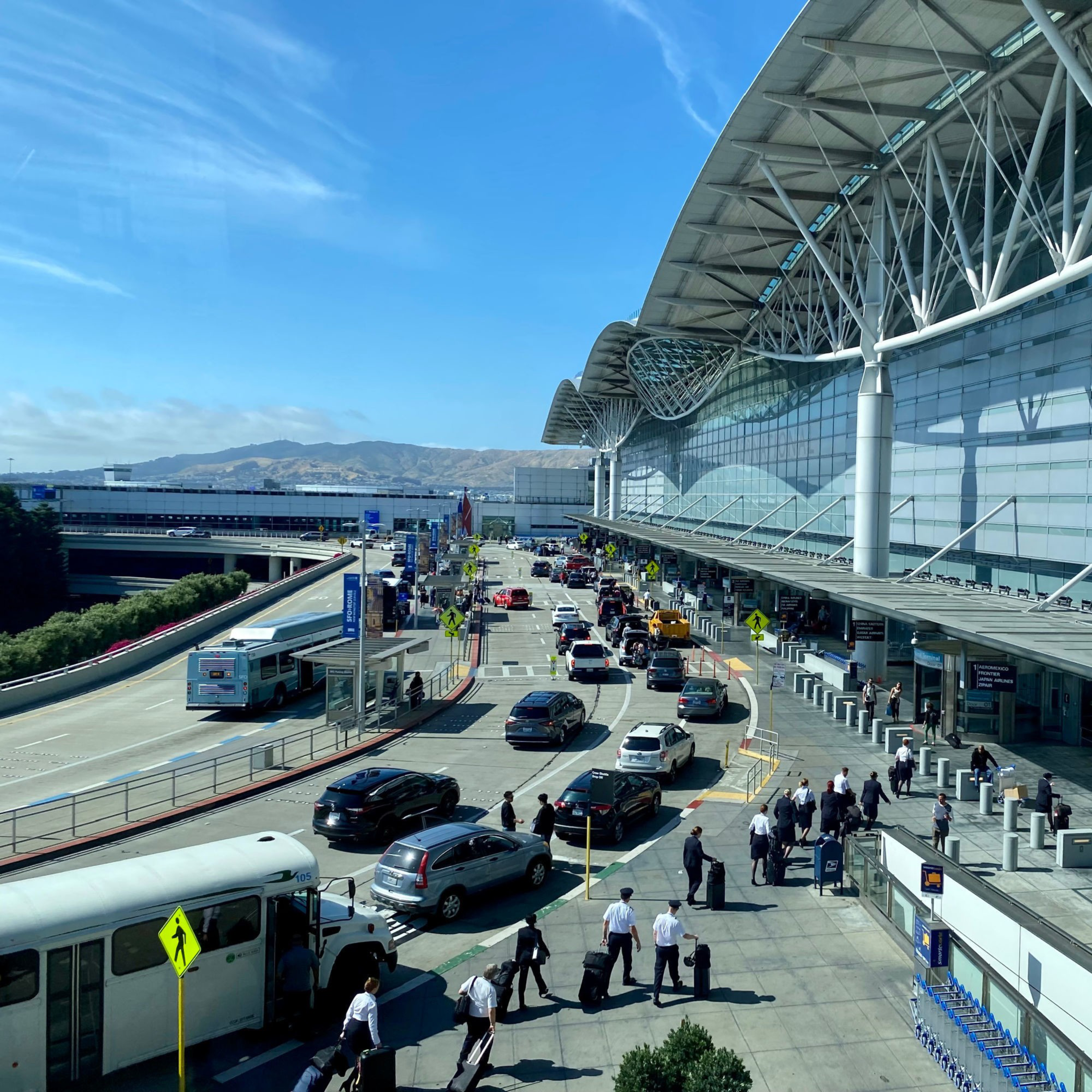 Cars and buses line up outside a modern airport terminal with large glass windows, while people walk with luggage under a clear blue sky.