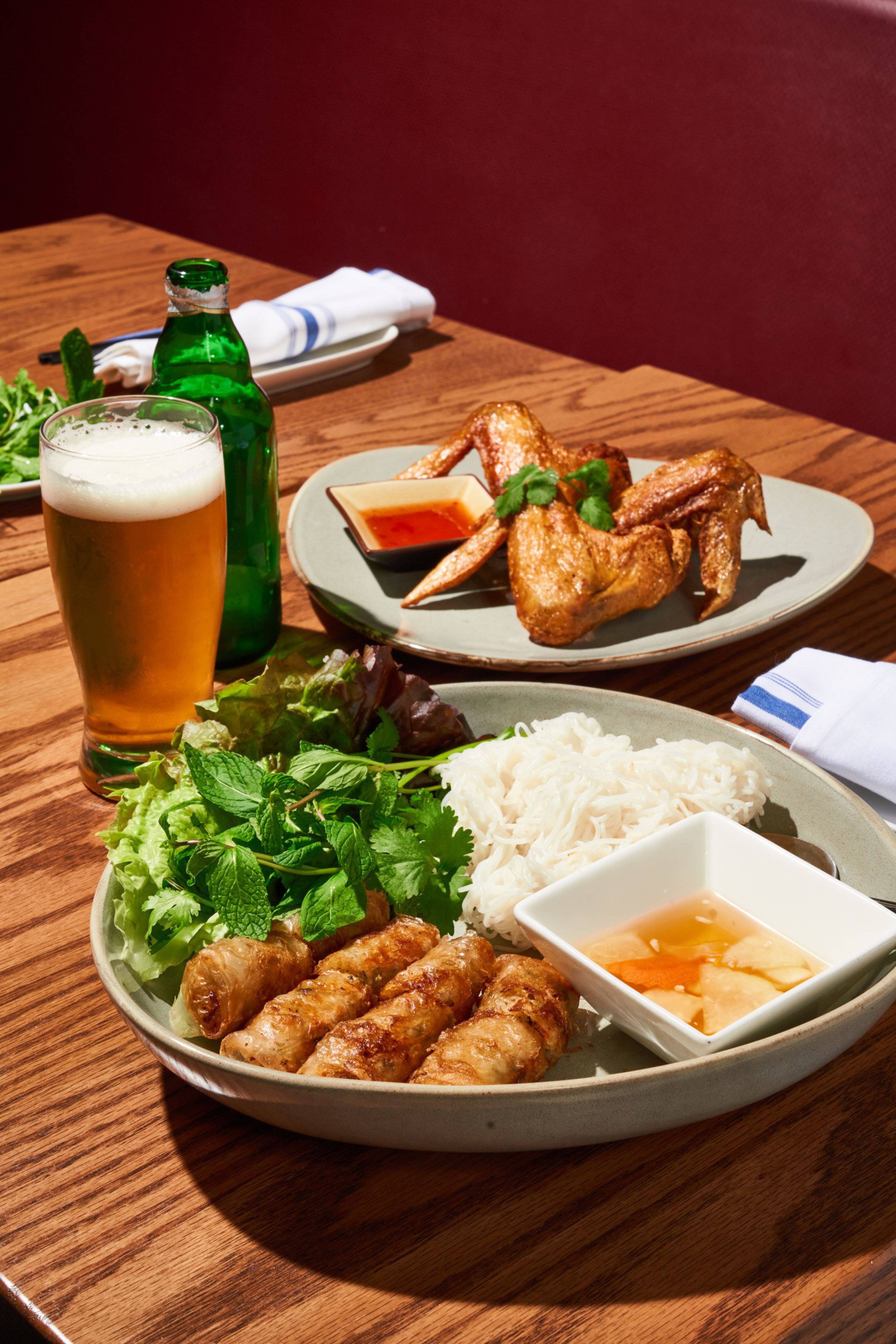 A table with a plate of spring rolls, fresh greens, noodles, dipping sauce, chicken wings, a green bottle, and a glass of beer, all set on a wooden surface.