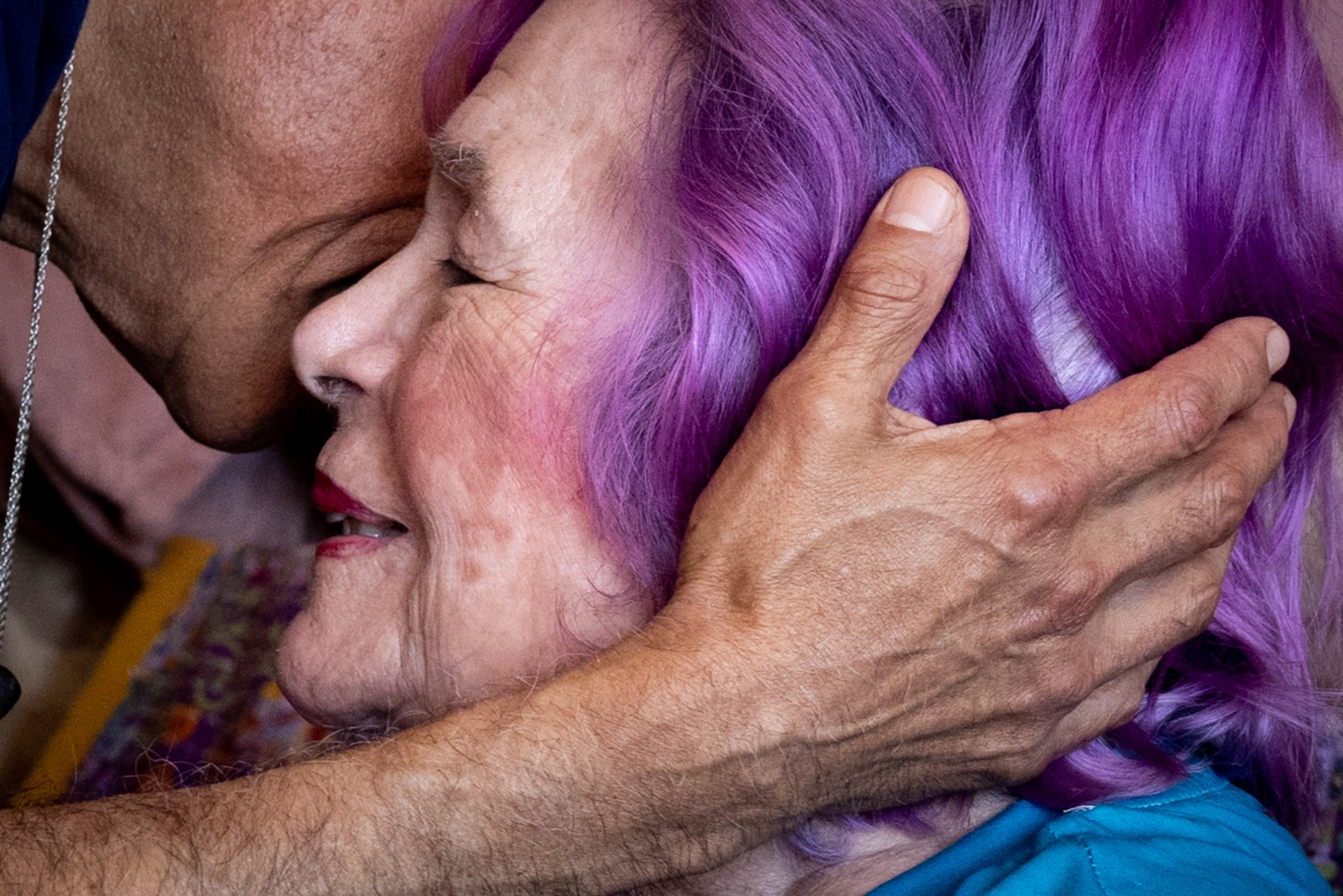 An elderly woman with vibrant purple hair is gently embraced and kissed on the forehead by a person with a tan arm, expressing tenderness.