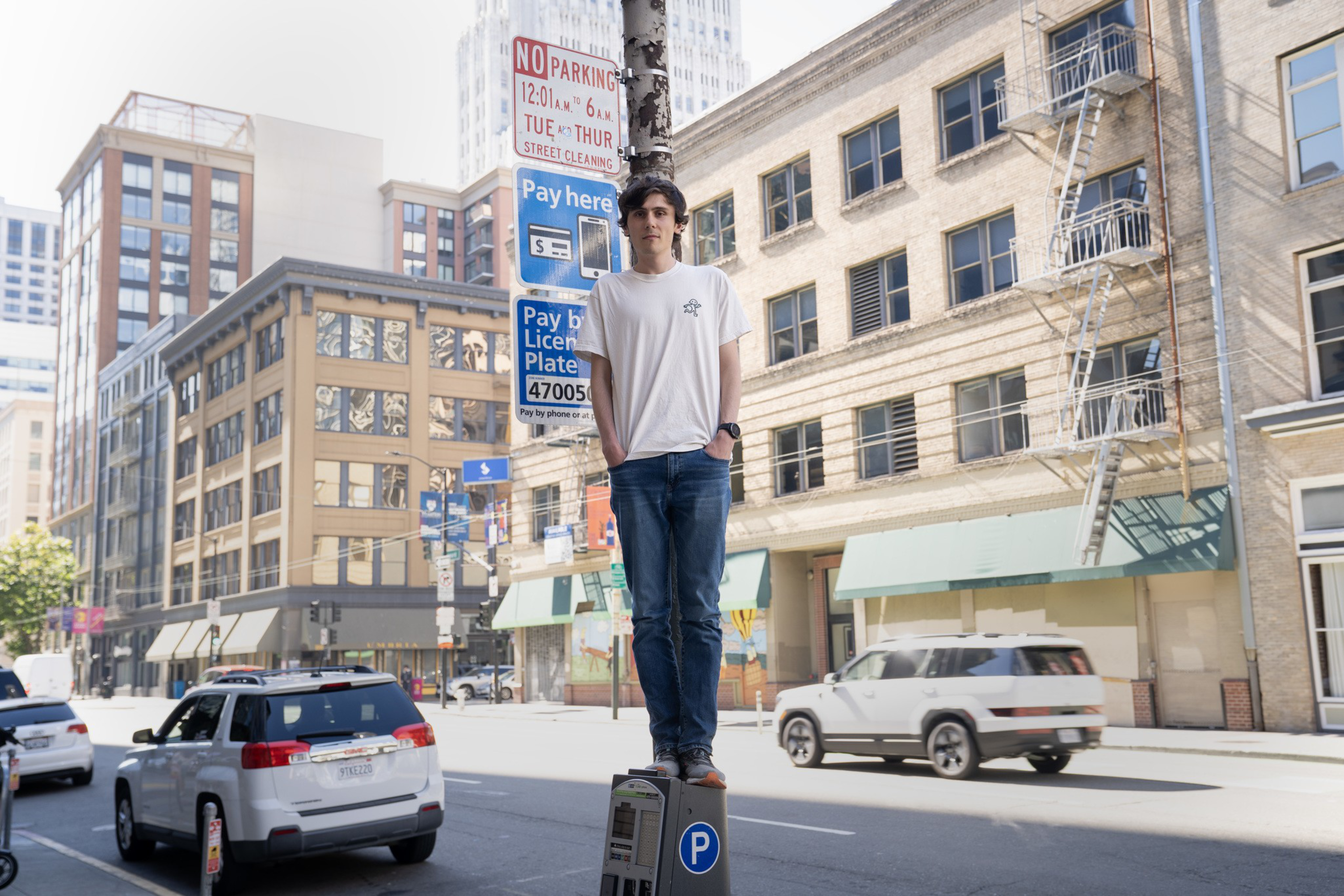 A young man in a white t-shirt and jeans stands on a parking meter on a city street with buildings, cars, and parking signs around him.