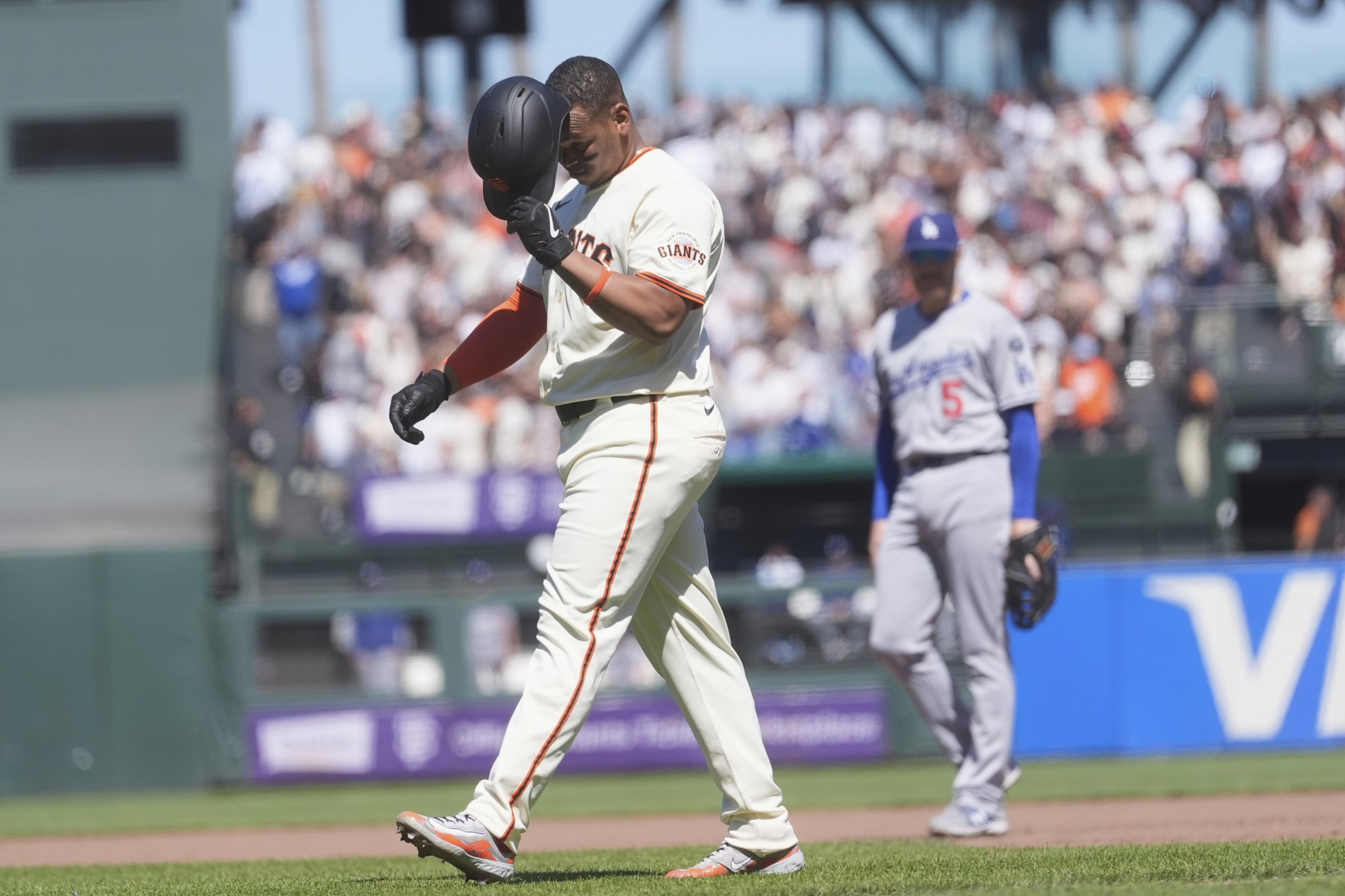A baseball player in a Giants uniform walks on the field, holding his helmet. A player in a gray Dodgers uniform stands in the background. A crowd is watching.