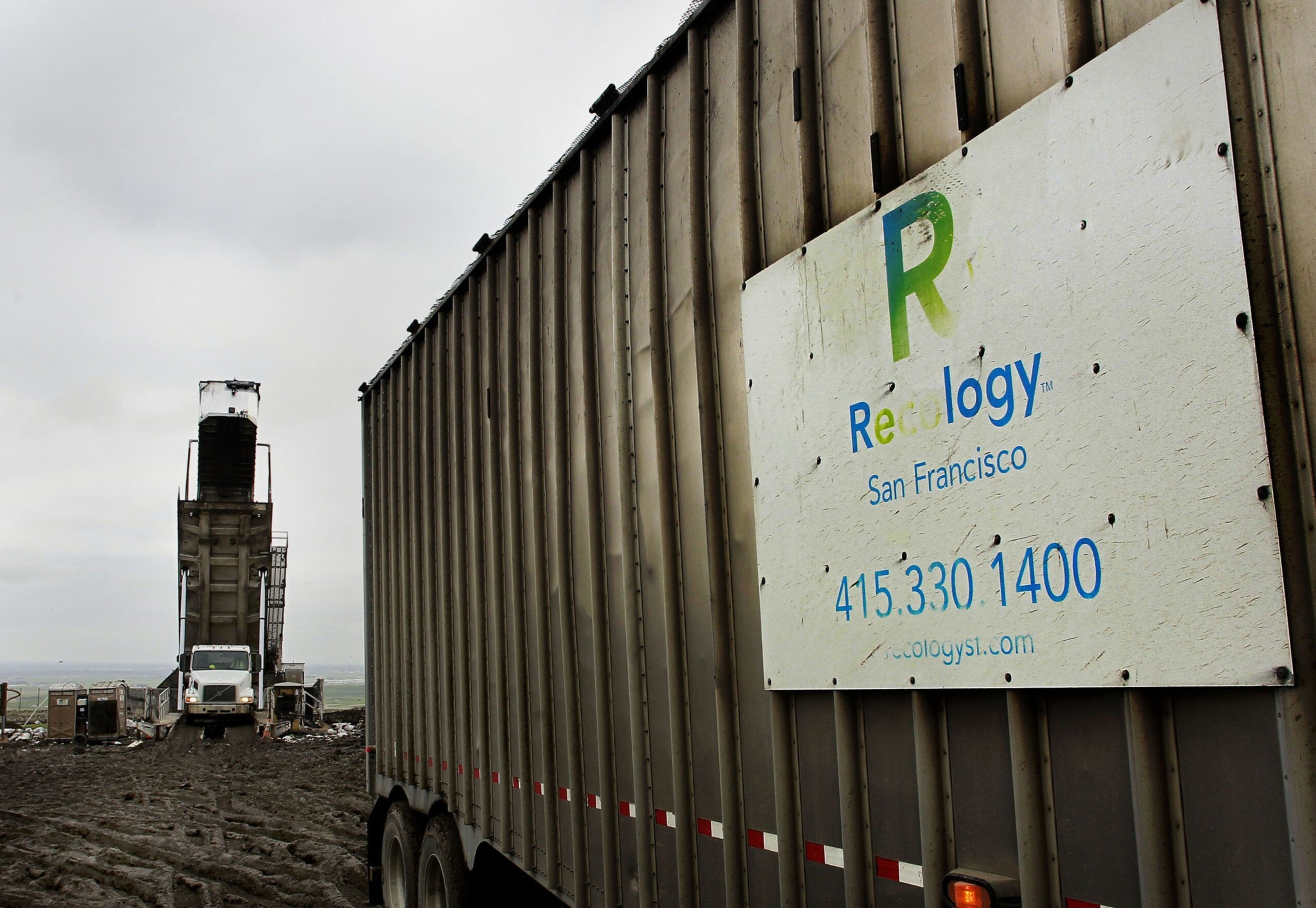 A large industrial container with a "Recology San Francisco" sign and phone number sits on muddy ground near a truck unloading waste into a tall processing tower.