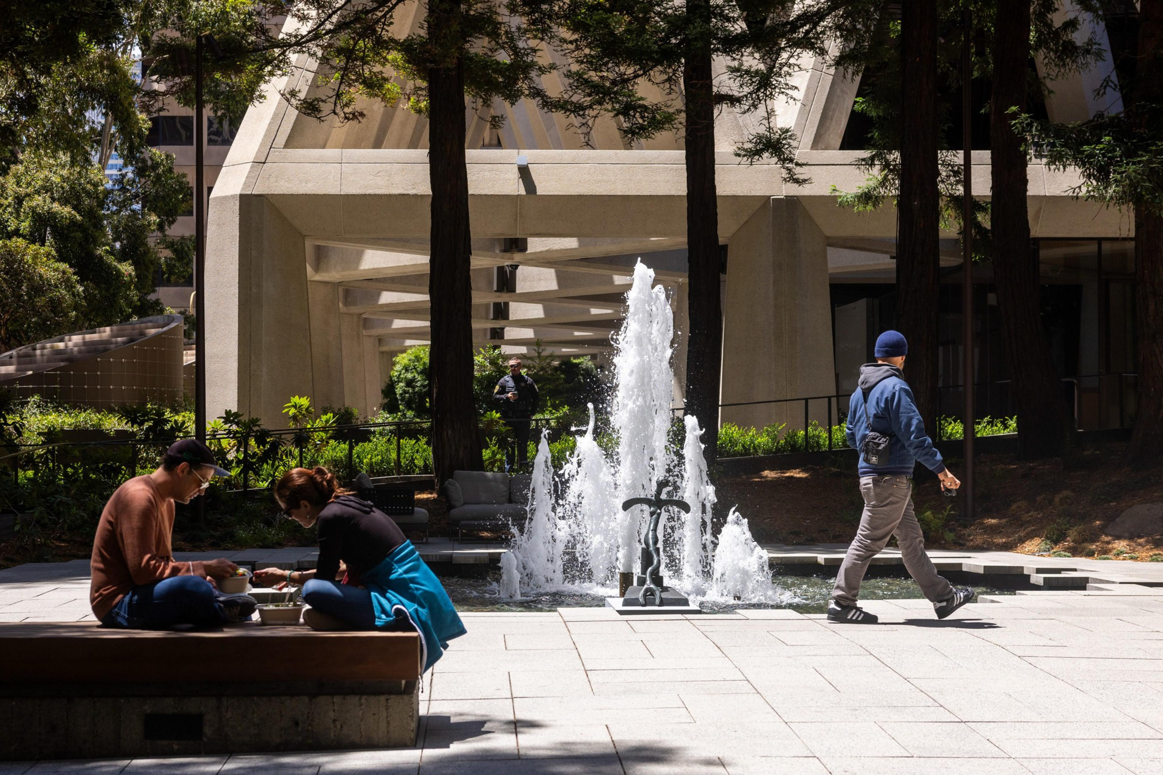 A couple sits on a bench eating, near a fountain with water jets. A person walks past, and the scene is framed by trees and a modern, concrete structure in the background.