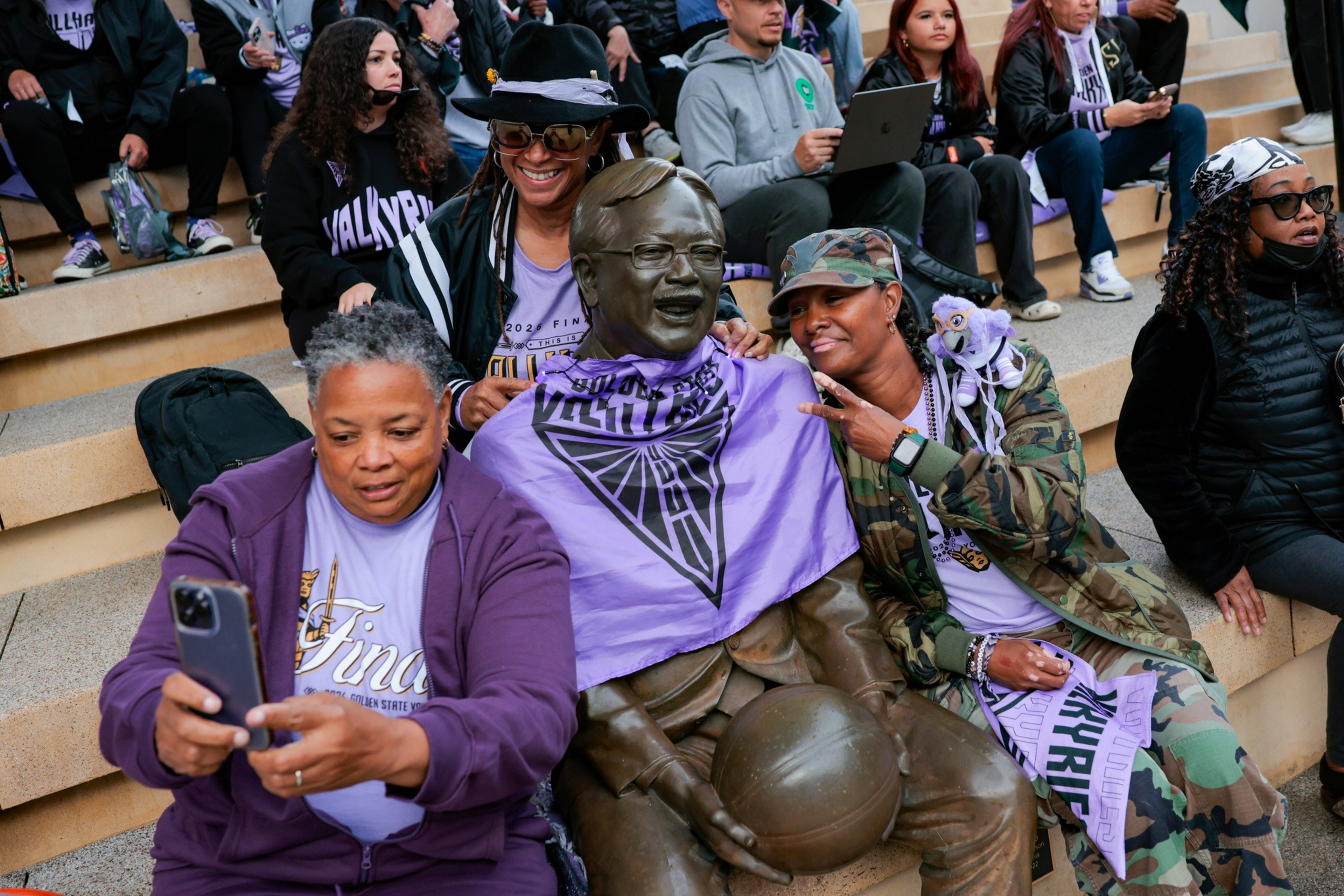 Three women sit on steps with a bronze statue of a man holding a basketball, smiling and taking a selfie while wearing purple fan apparel.