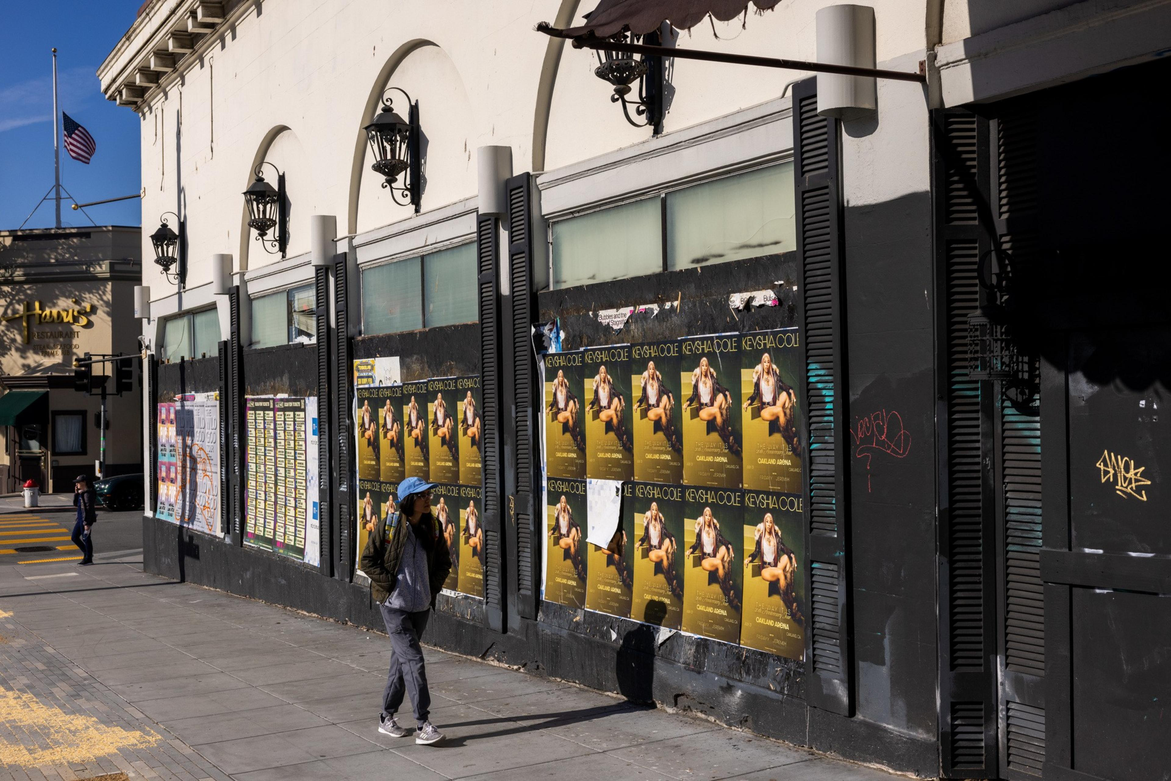 A person walks past a building with black shutters and multiple posters of the same image plastered on its wall. An American flag is seen in the background.