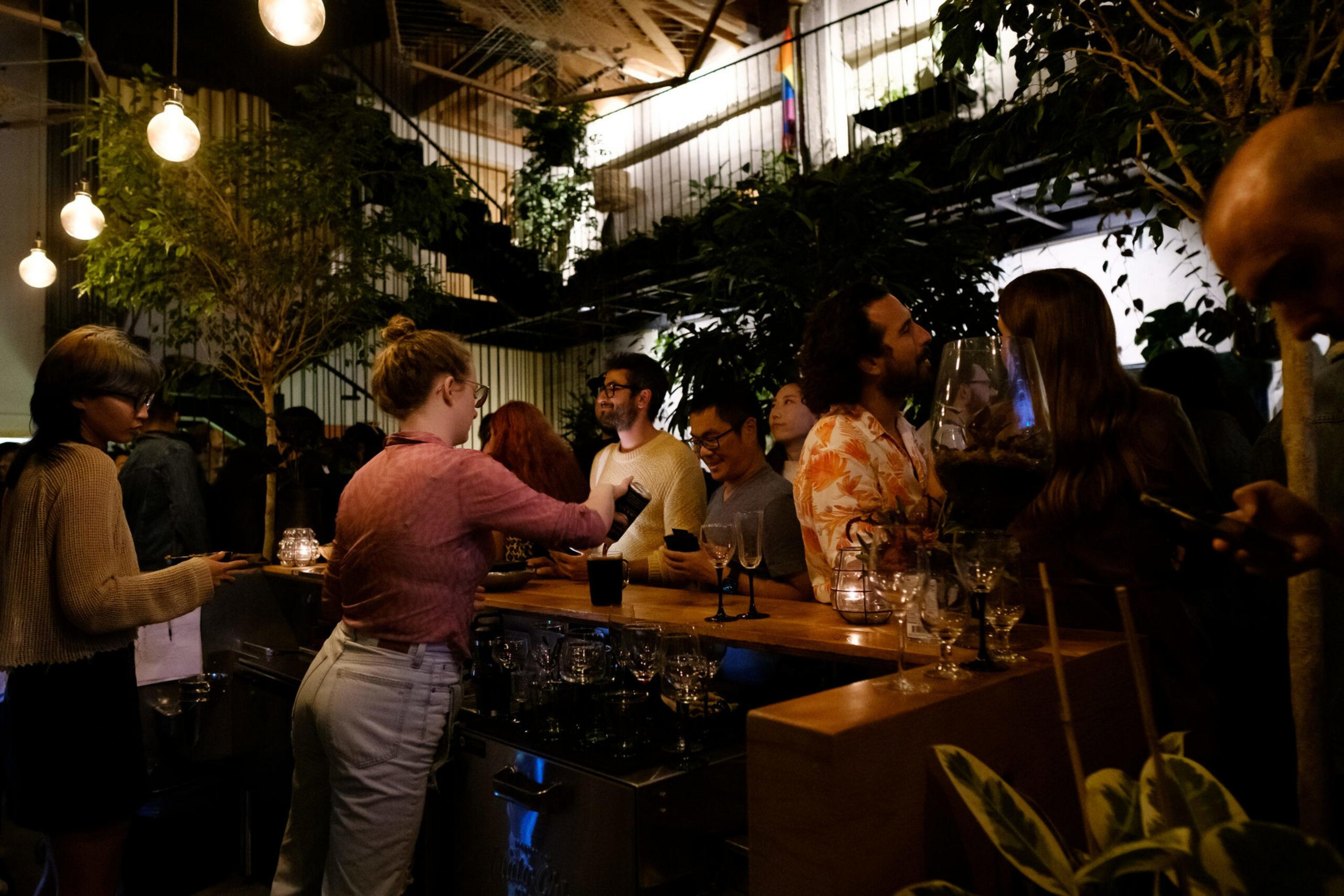 People gather around a bar lit by warm hanging bulbs and surrounded by lush greenery. They chat and sip drinks, creating a cozy, lively atmosphere.