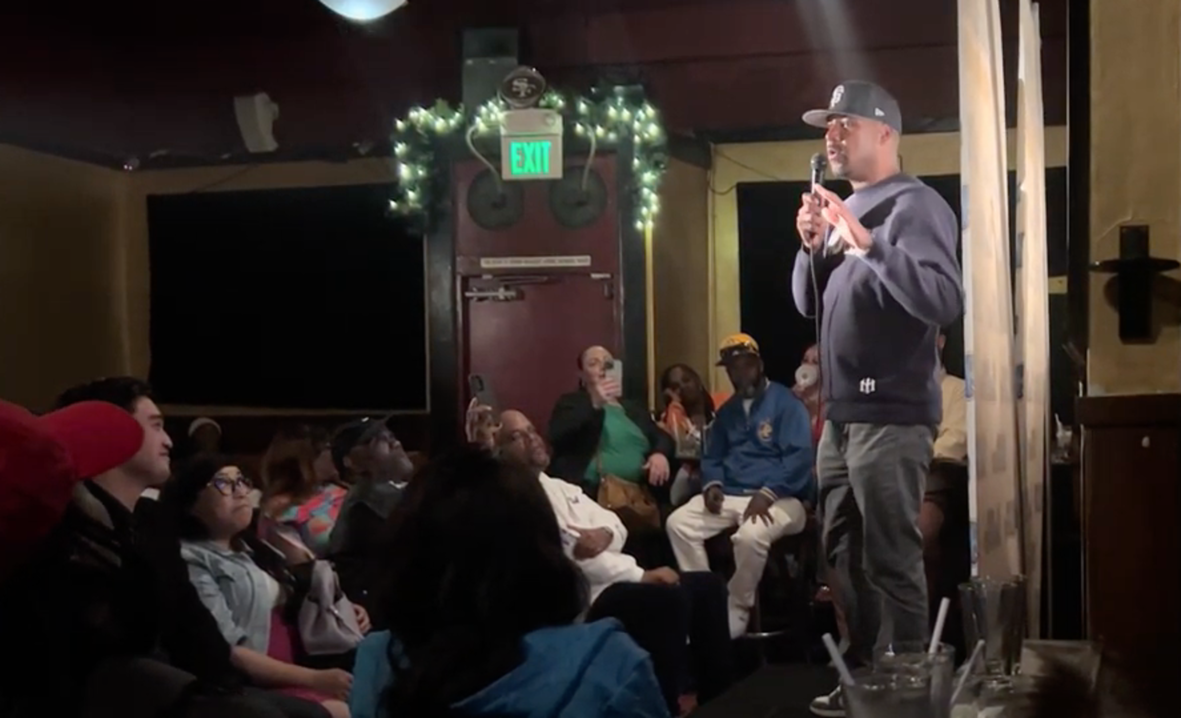 A man wearing a cap and sweater speaks into a microphone on a small stage as a seated audience listens attentively in a dimly lit room.