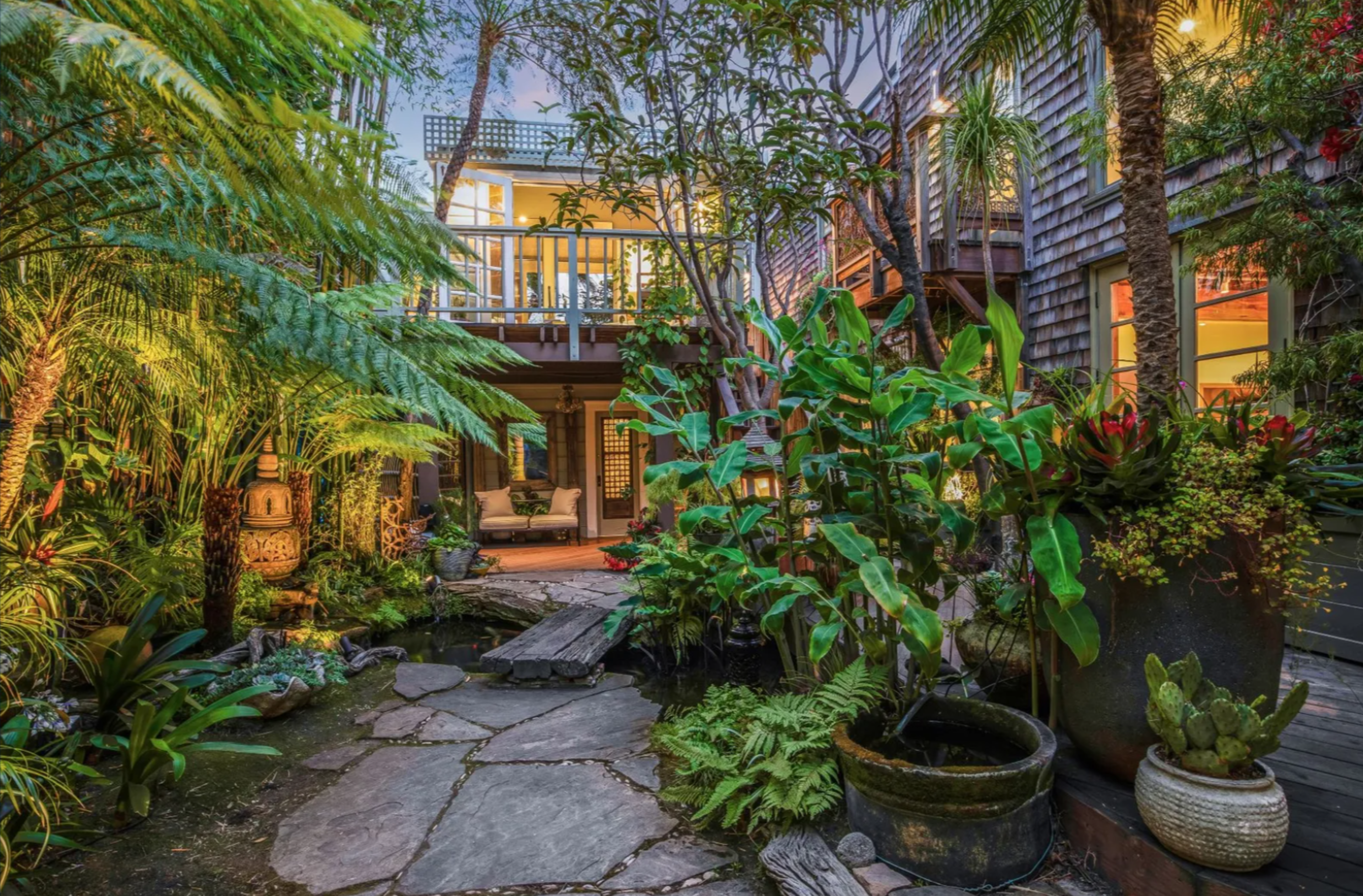 A lush garden courtyard with stone pathways, various plants in pots, a small wooden bridge over a pond, and a cozy patio area surrounded by house walls.