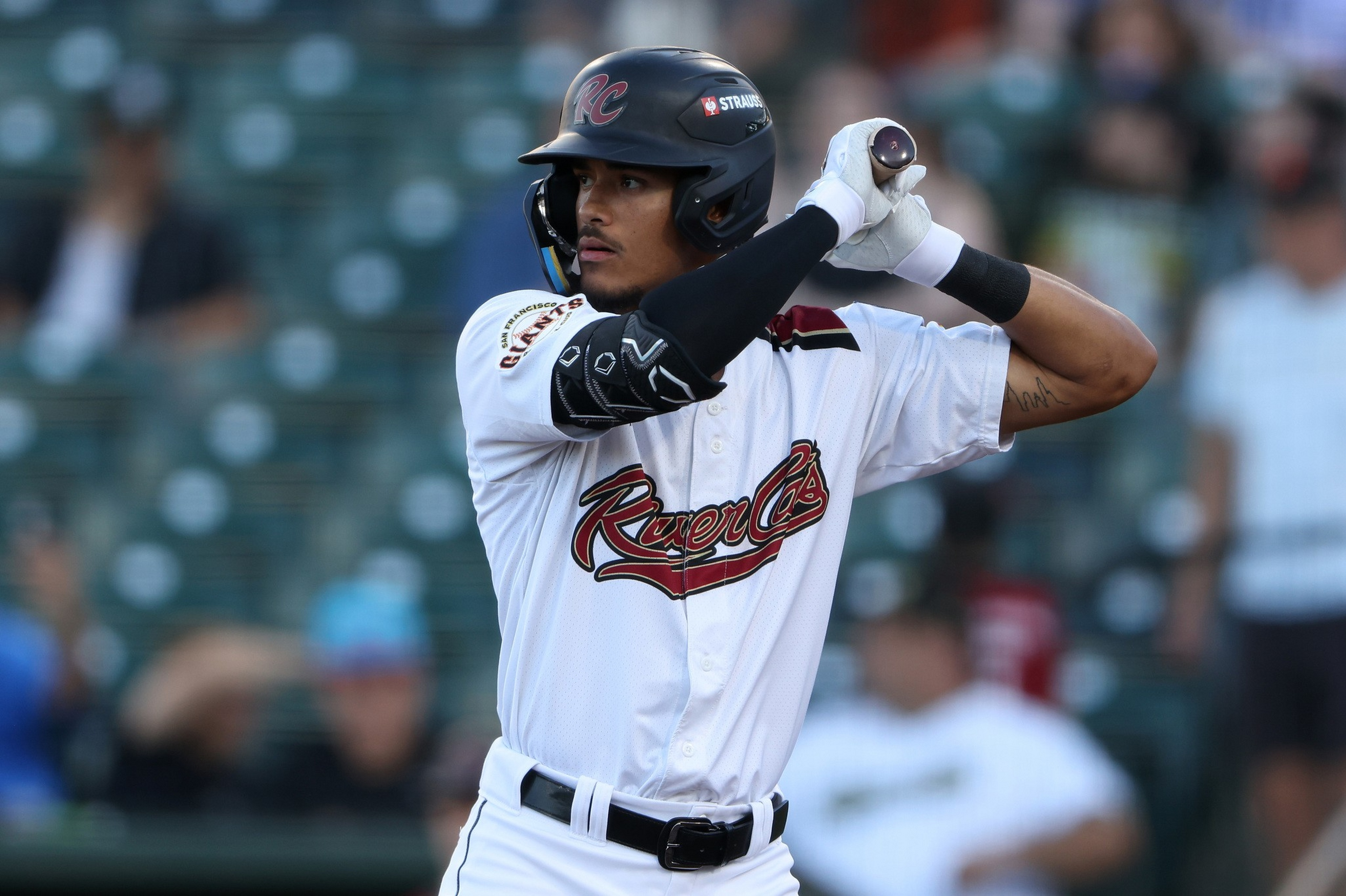 A baseball player in a white River Cats uniform and black helmet grips a bat, preparing to swing during a game.