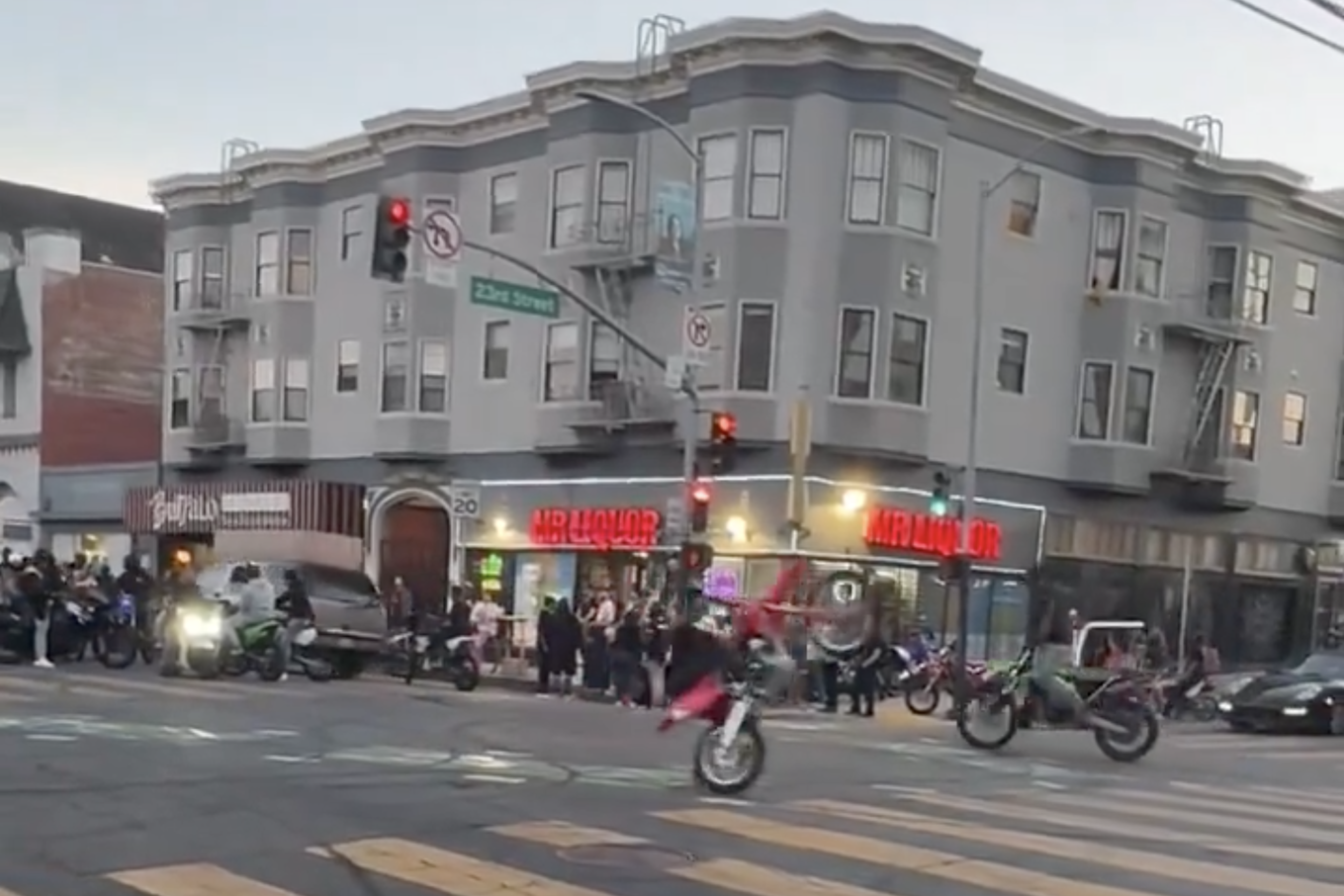 A bustling street corner with people gathered, motorcycle riders performing stunts, a grey building with a bar and liquor store, and traffic lights glowing red.
