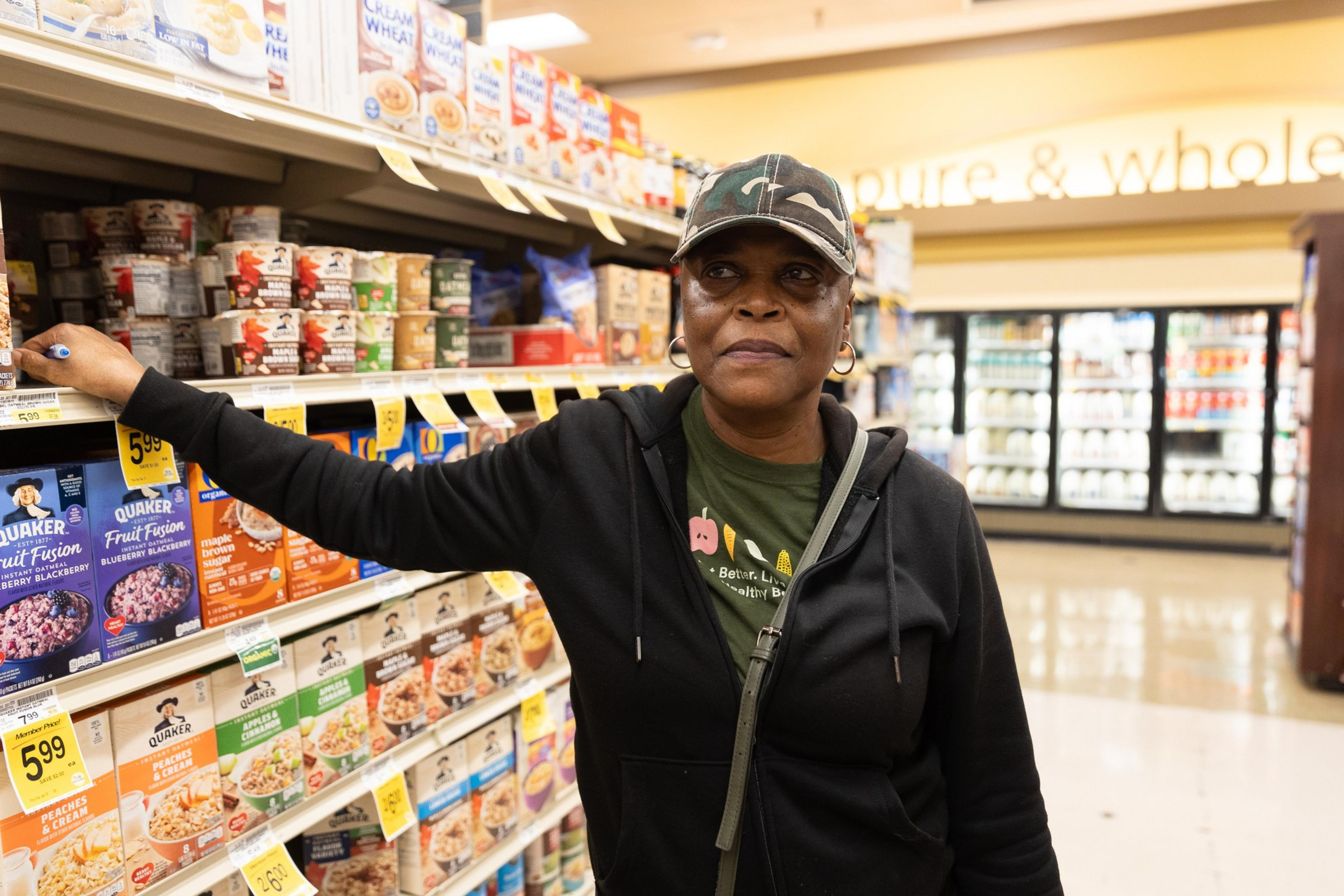 A person in a black hoodie and camo cap stands in a grocery aisle, surrounded by cereal boxes. Their hand rests on a shelf as they look ahead.