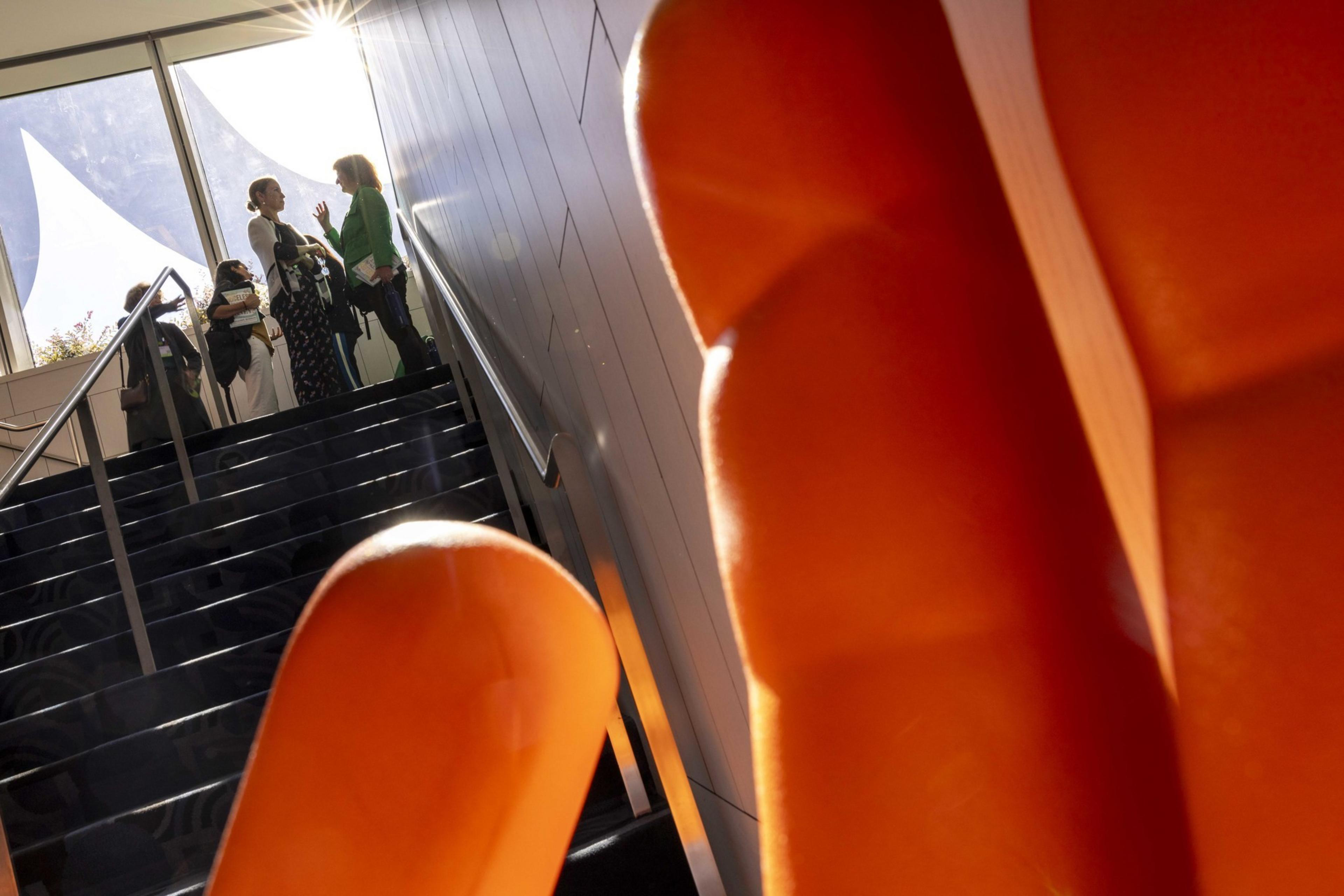 Two people are talking at the top of a staircase bathed in sunlight, with orange abstract shapes in the foreground and bright windows in the background.