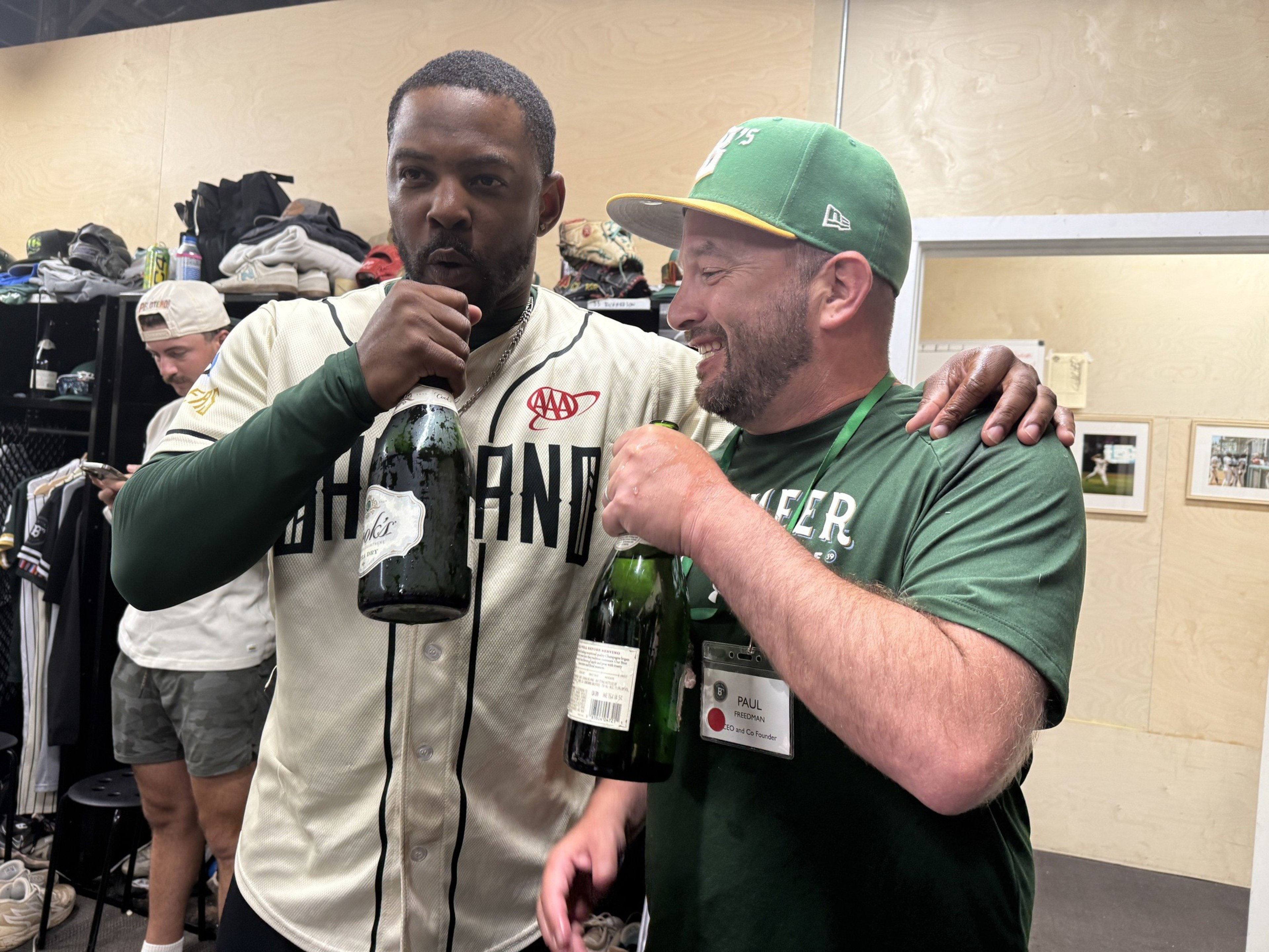 Two men cheer with champagne bottles in a locker room; one wears a Cleveland baseball jersey, the other a green shirt and cap with a "Paul" name tag.