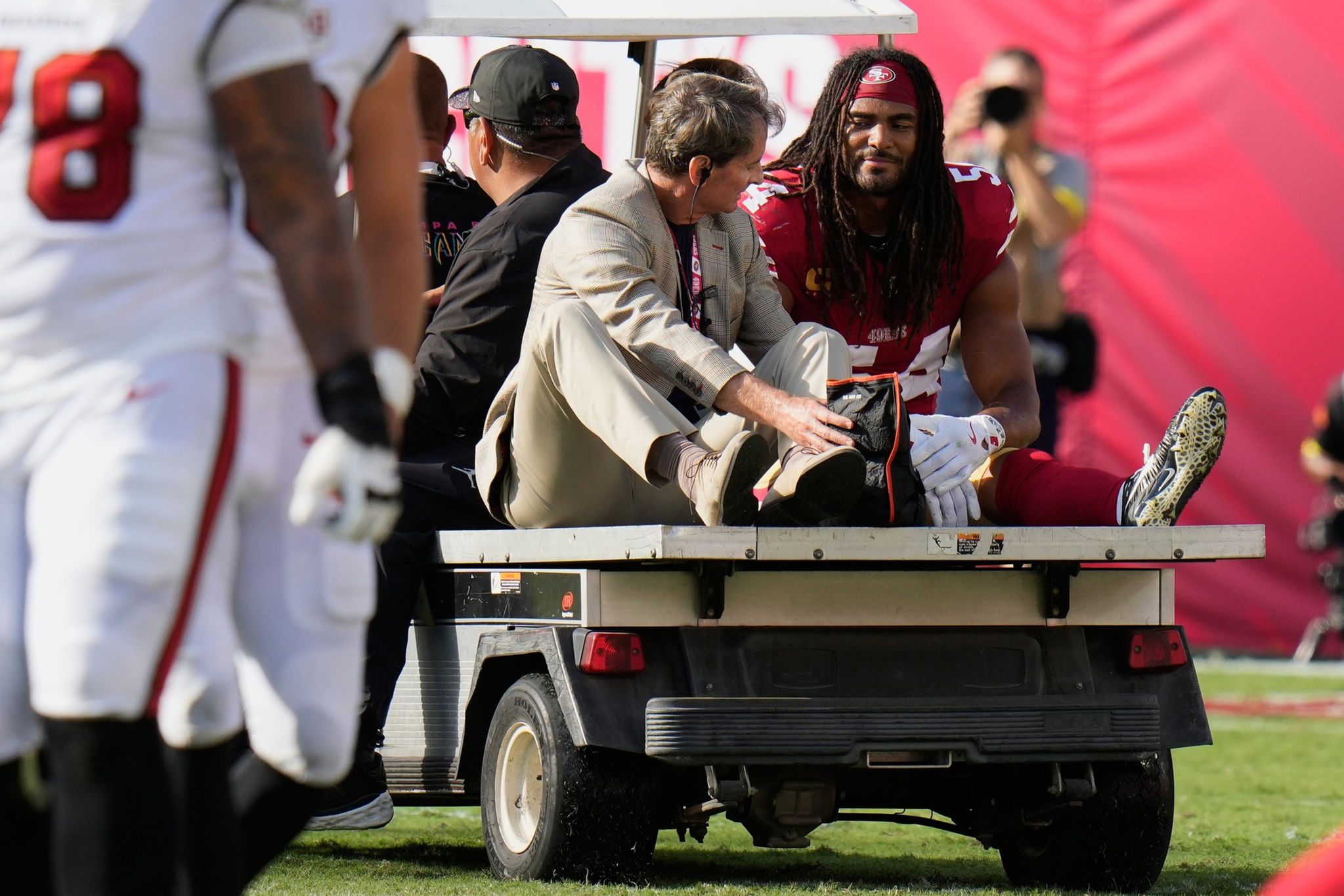 A football player in a red uniform sits on a golf cart receiving medical attention from a man in a beige jacket, while other players stand nearby.