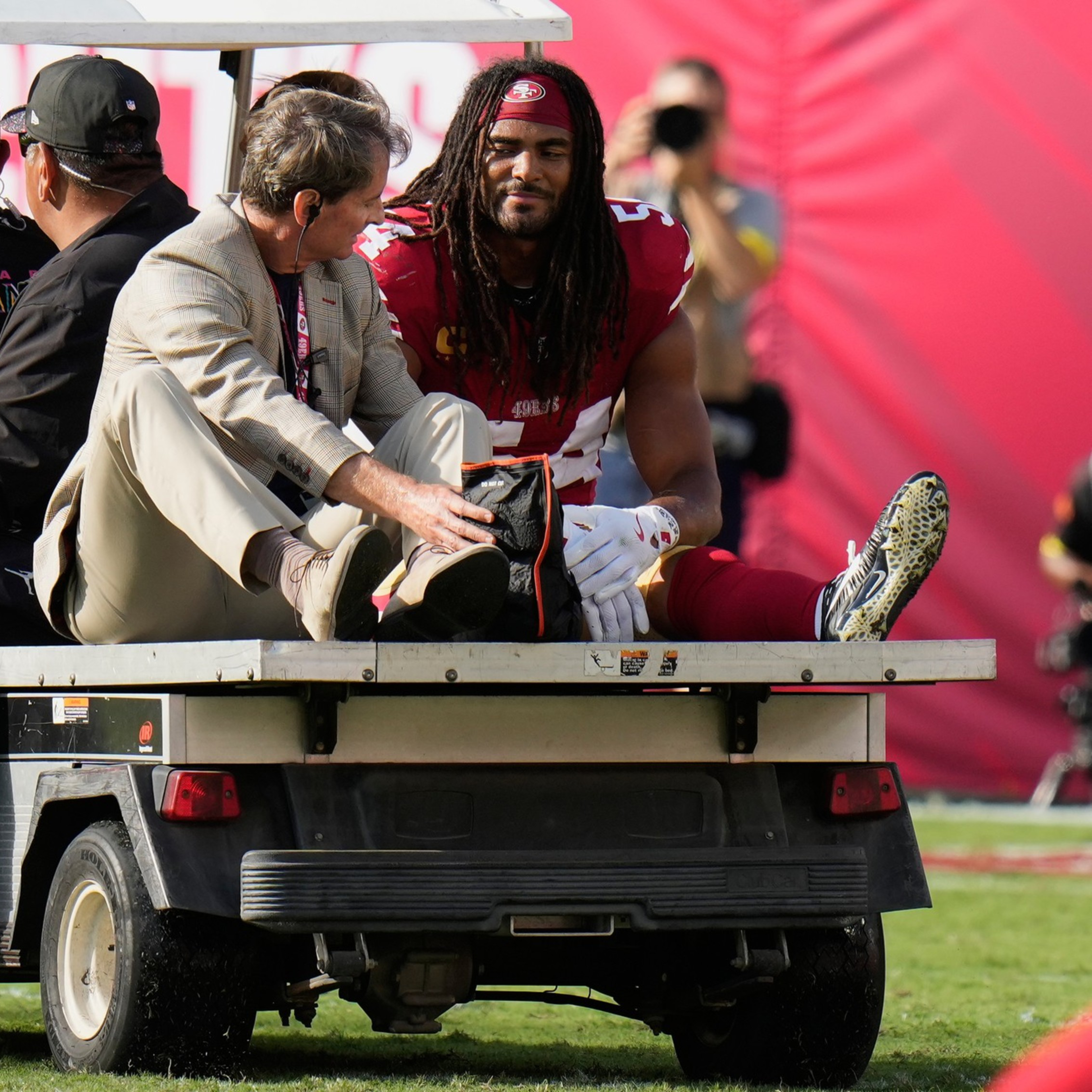 A football player in a red uniform sits on a golf cart receiving medical attention from a man in a beige jacket, while other players stand nearby.
