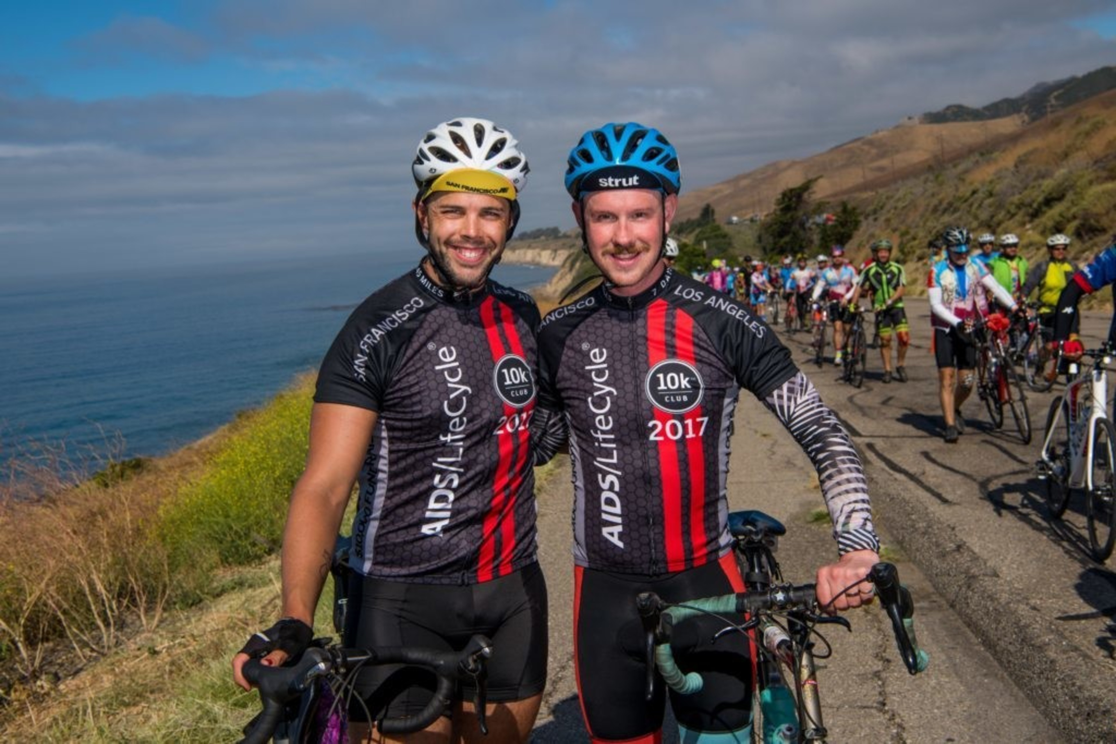 Two cyclists in matching jerseys and helmets stand smiling with their bikes on a scenic coastal road, while more cyclists are seen in the background.