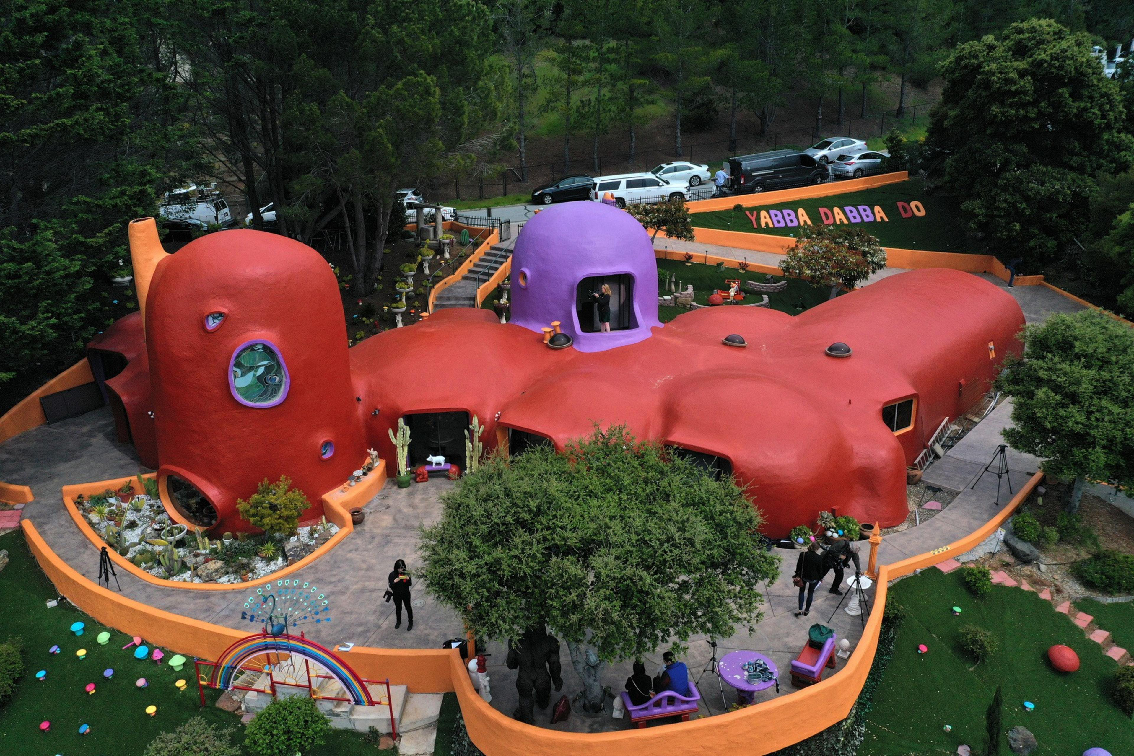 The image shows a colorful, whimsical house resembling The Flintstones' home, with vibrant orange and purple domes and surrounded by lush green landscape.