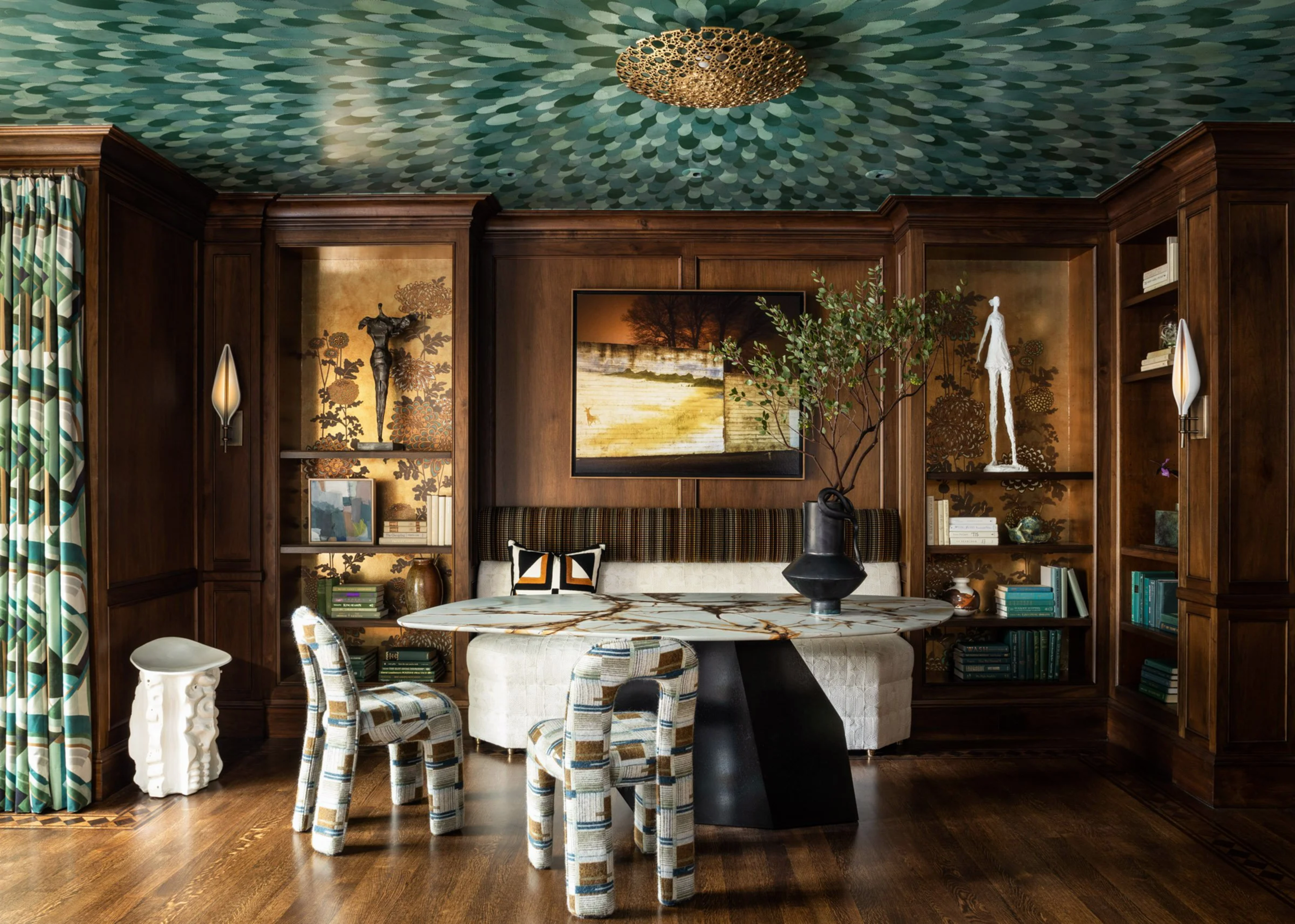 A cozy room features dark wood paneling, patterned blue-green ceiling, a marble table with curved legs, two patterned chairs, and a leafy plant centerpiece.