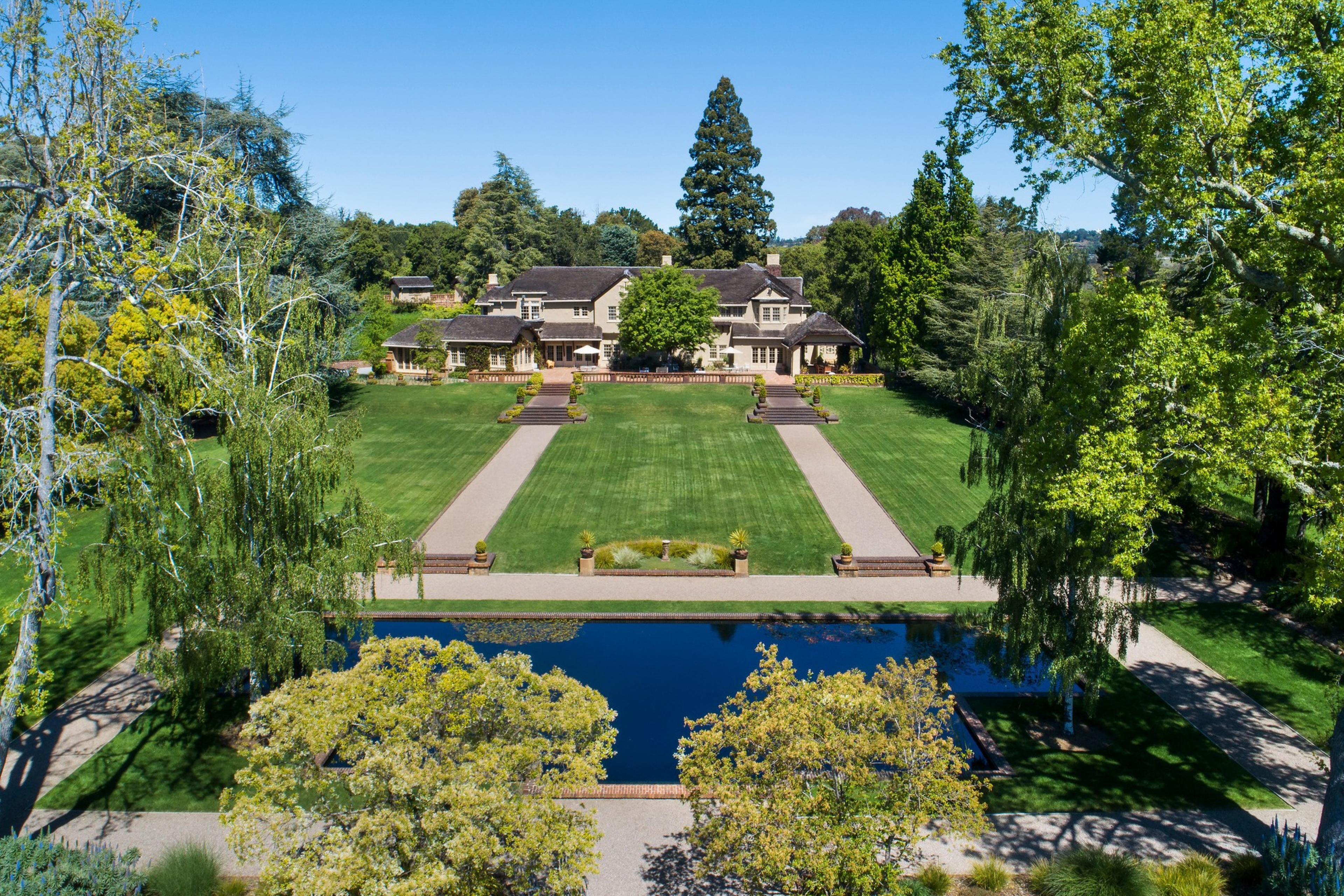 A large mansion sits behind a wide, manicured lawn with two parallel paths leading to it, bordered by tall trees and a reflective rectangular pond in front.
