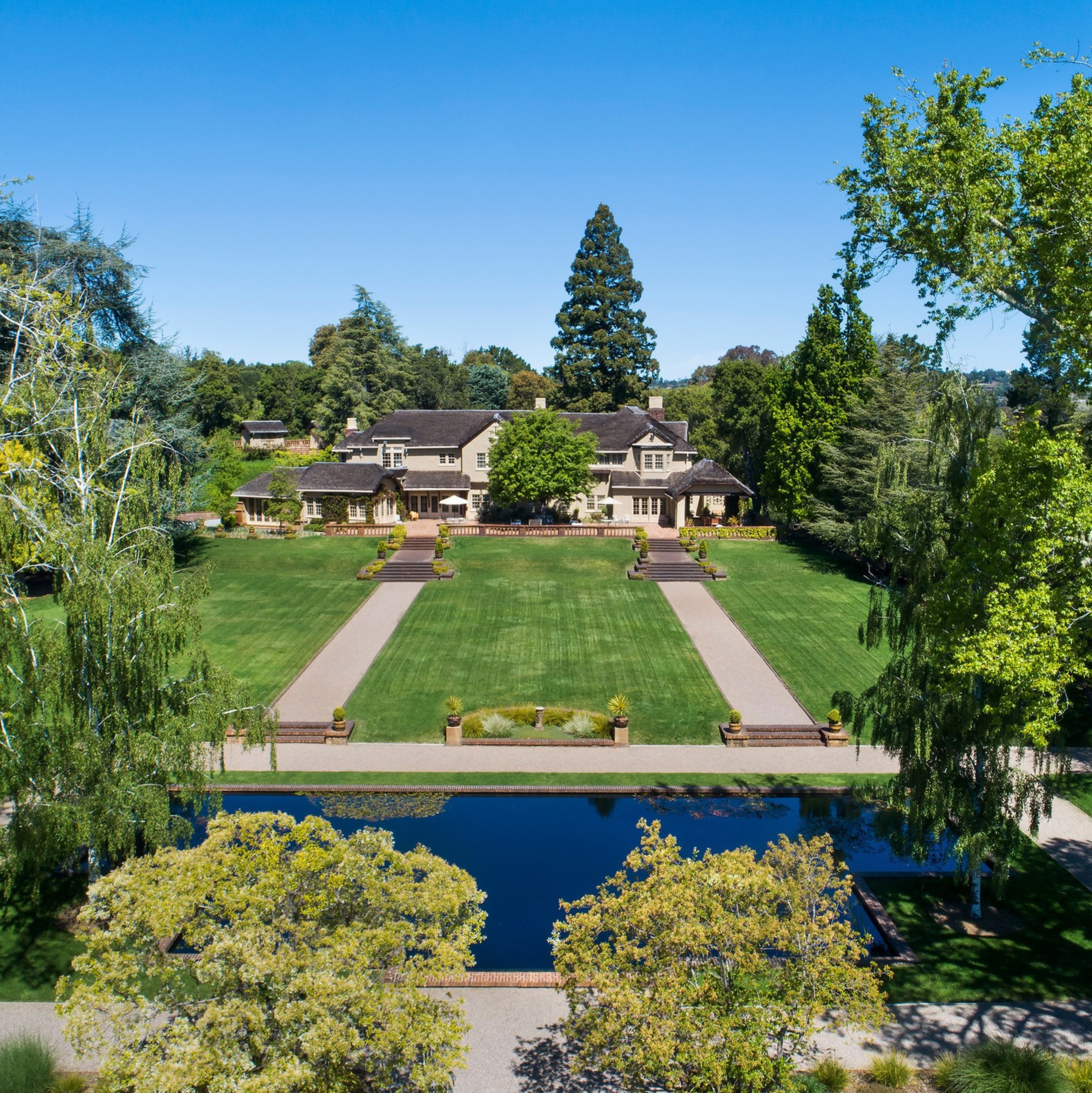 A large mansion sits behind a wide, manicured lawn with two parallel paths leading to it, bordered by tall trees and a reflective rectangular pond in front.