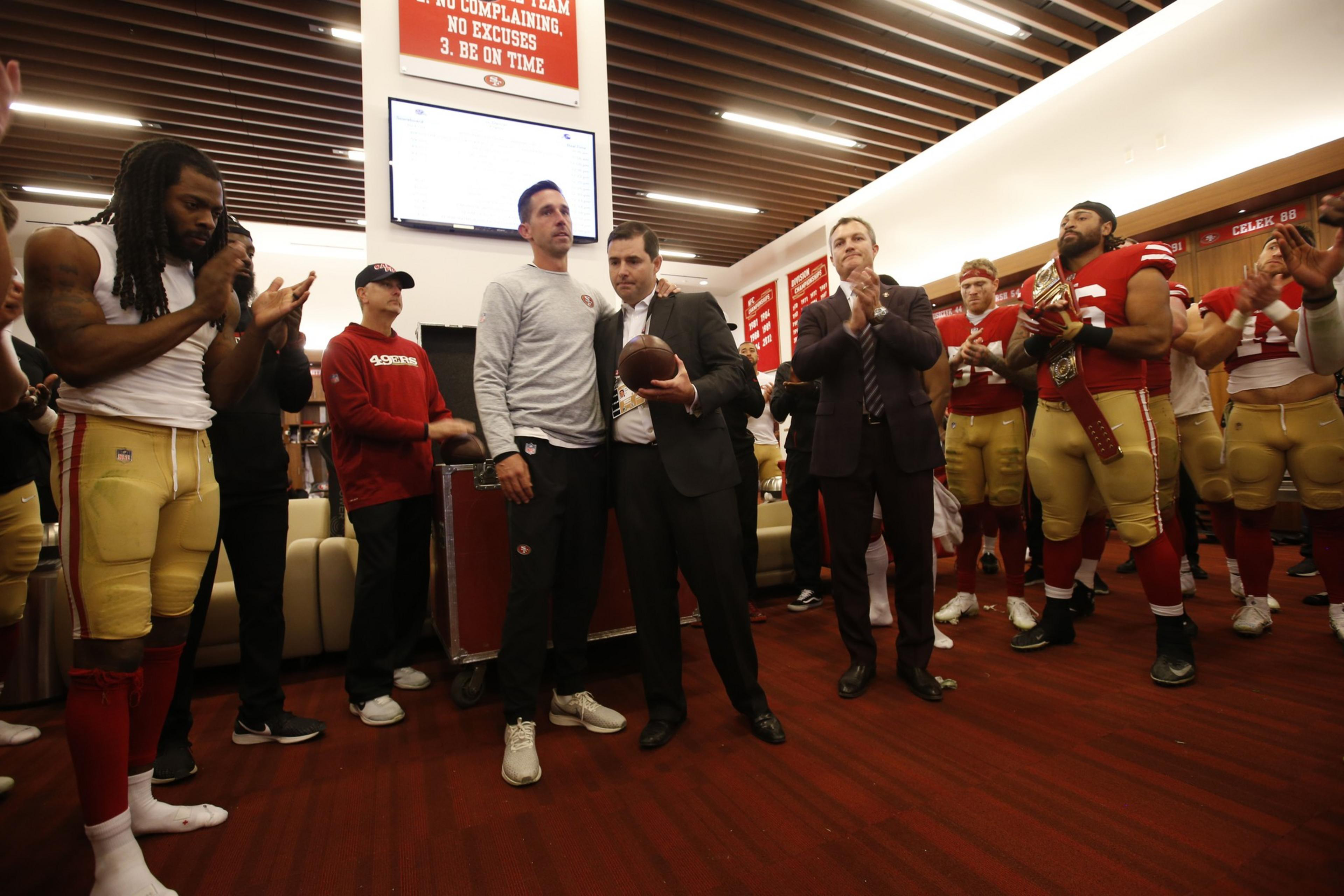 A group of people, including football players and a man in a suit, are gathered in a room. One person holds a football, while others are clapping.