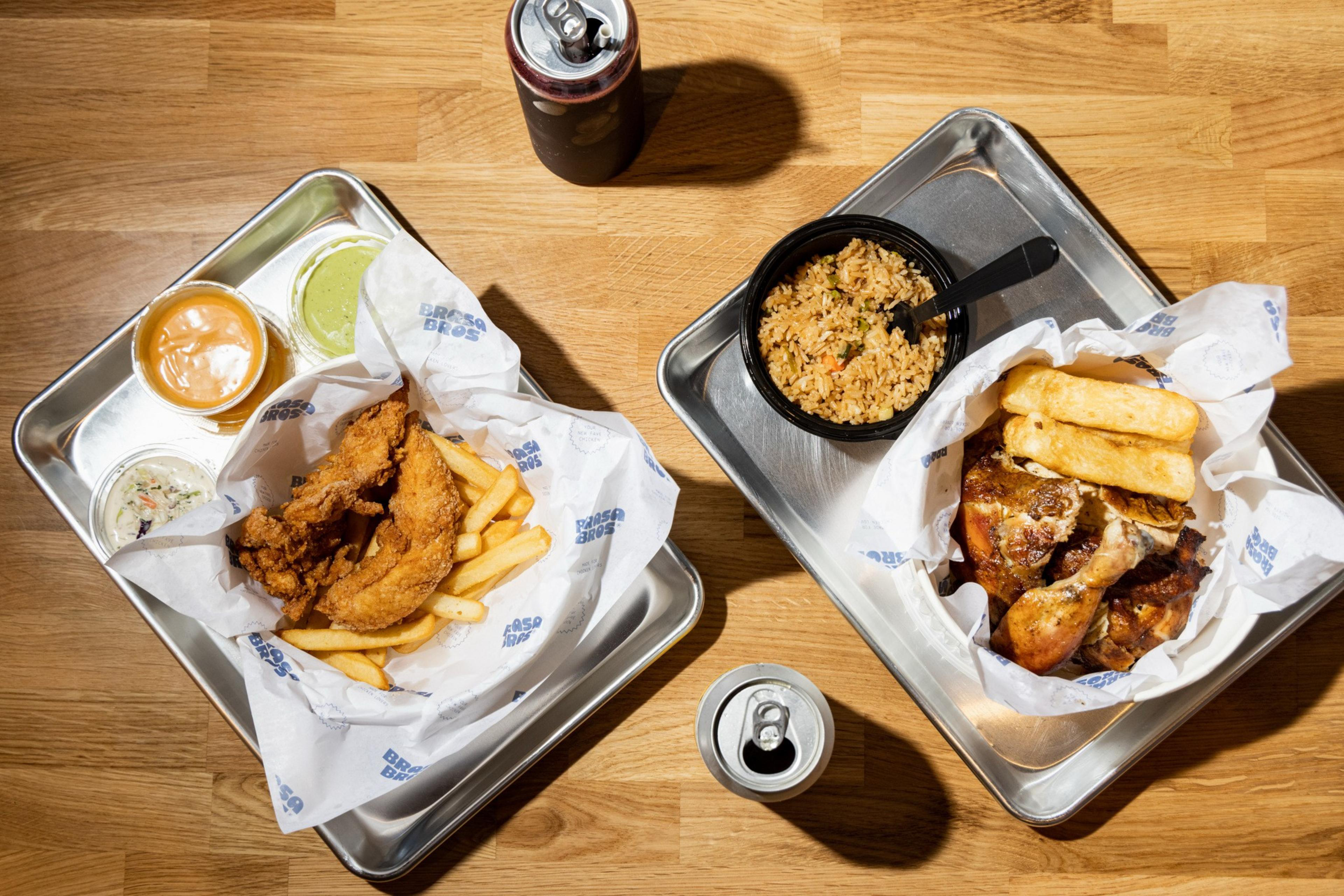 Two metal trays hold meals on a wooden table: one with fried chicken, fries, three sauces, and a soda can; the other with roasted chicken, boiled yuca, rice, and a soda can.