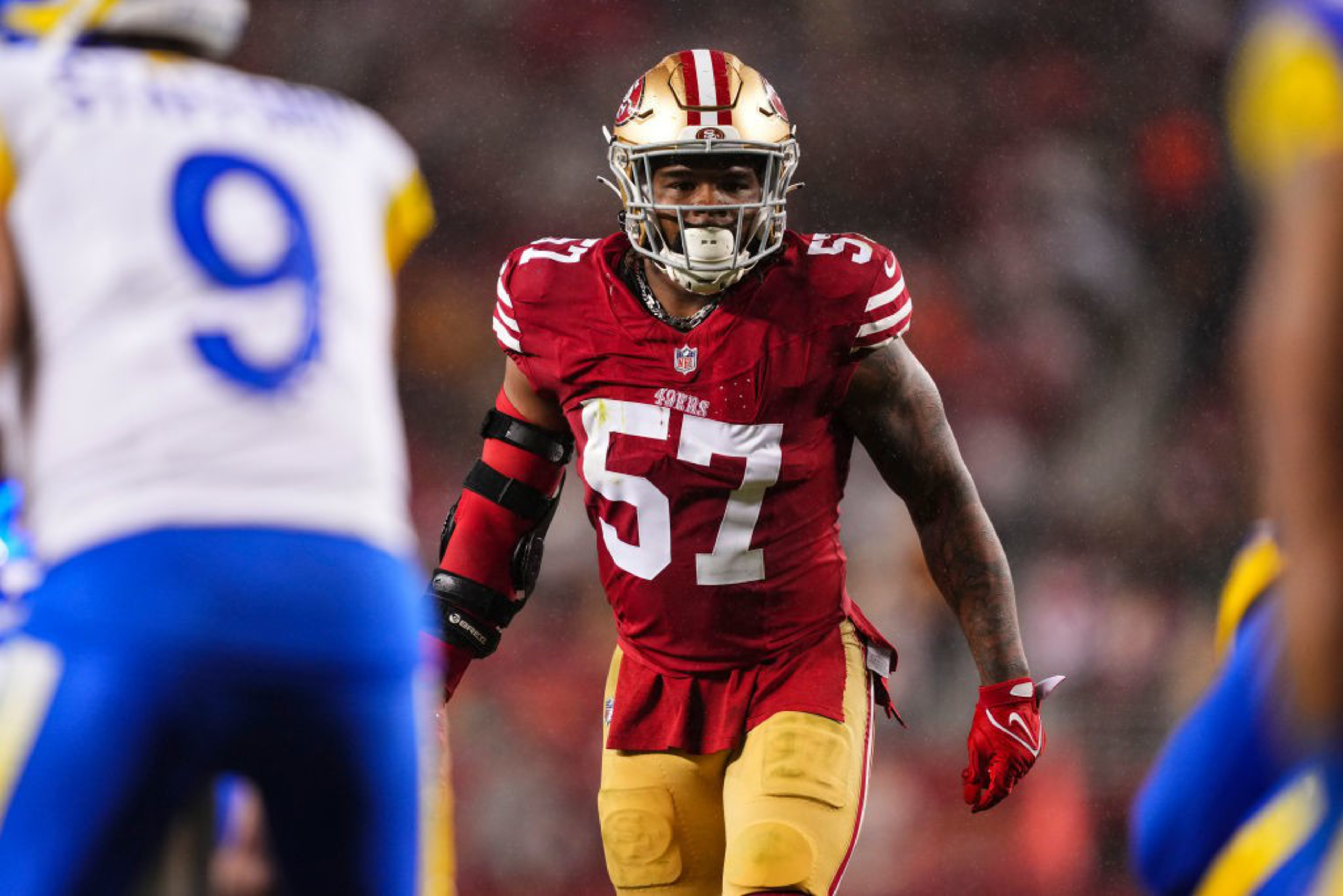 A football player in a red 49ers uniform with the number 57 is intensely focused on the field. Opponents in blue and white are partially visible.