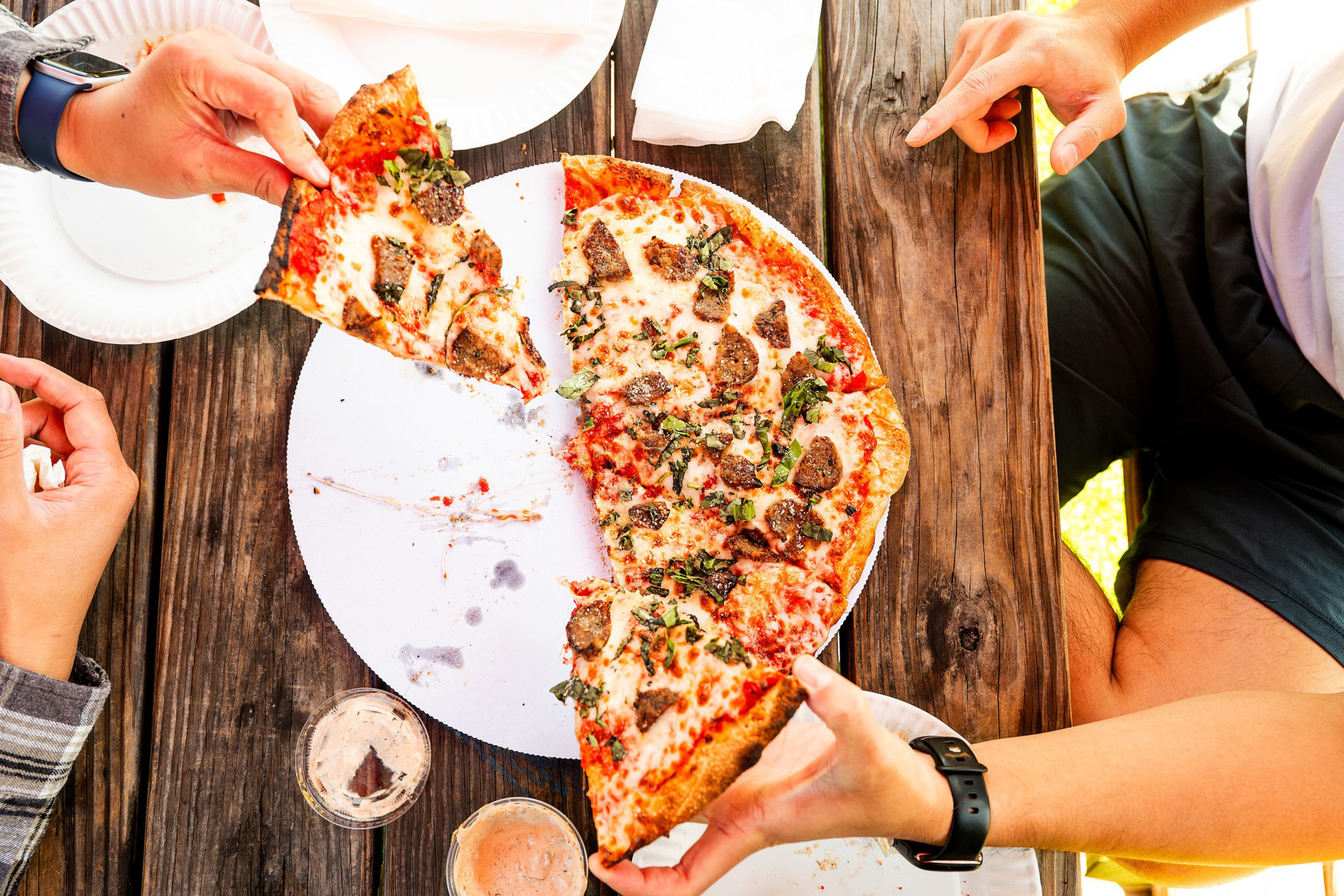Two people are sharing a large pizza with meat, cheese, and herbs on a rustic wooden table with drinks and paper plates.