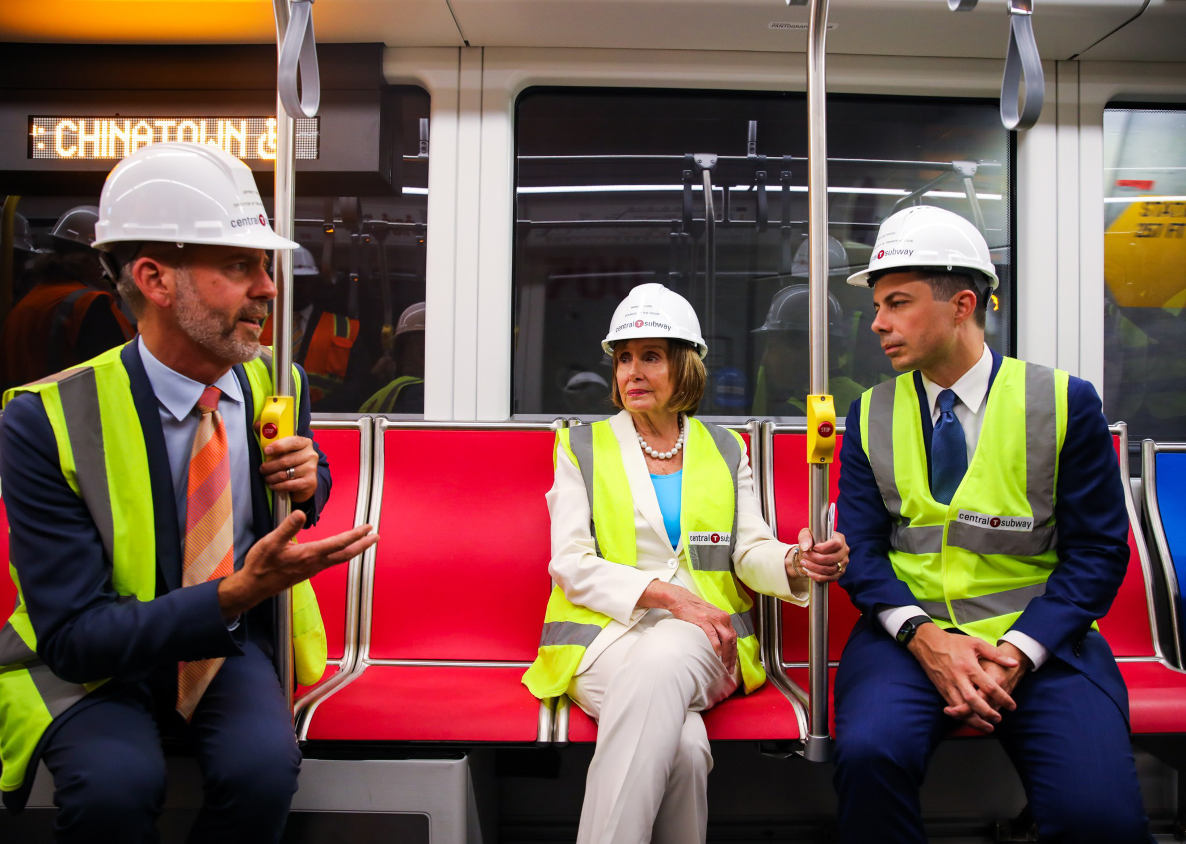 Three people wearing white hard hats and yellow safety vests sit on red seats inside a subway car, with one man talking and the others listening.