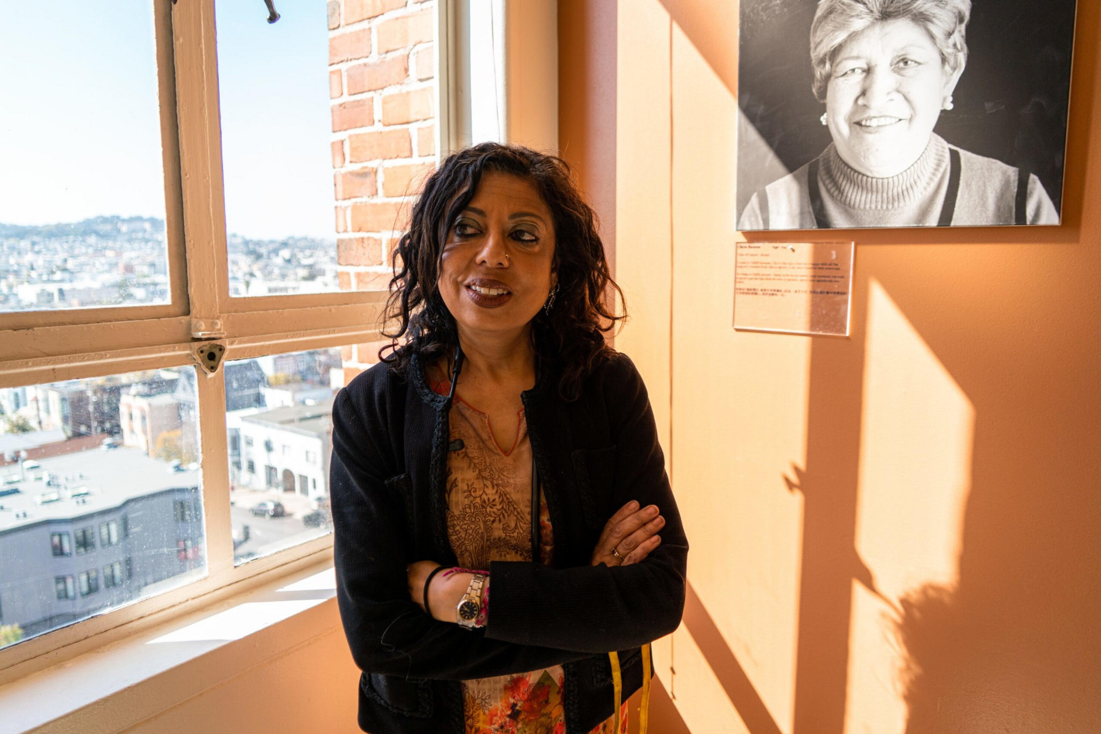 A woman with dark curly hair stands with arms crossed near a sunlit window, next to a wall displaying a black-and-white portrait of an older woman.