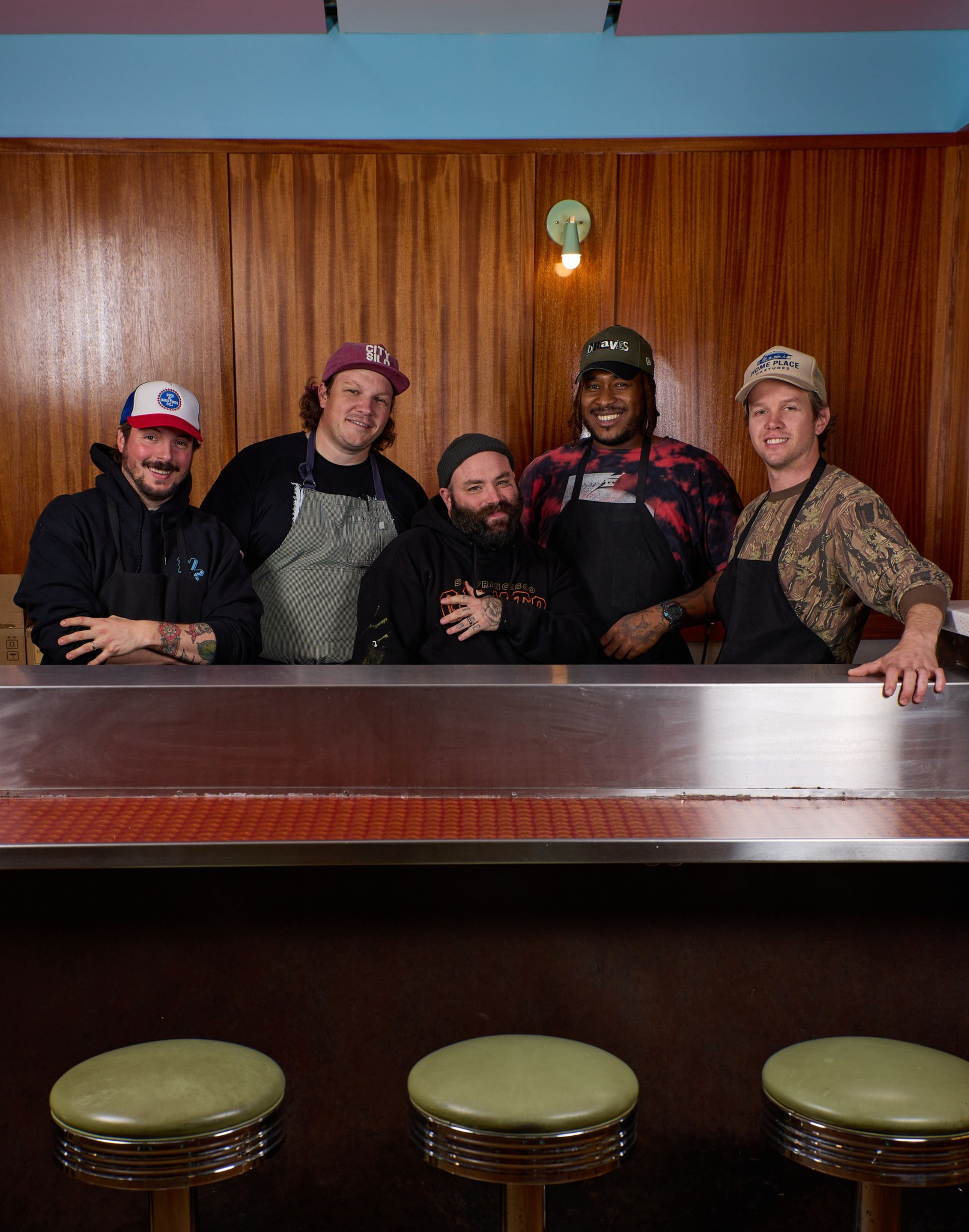Five men wearing aprons and casual hats stand behind a diner counter with three green stools, posing and smiling in front of a wood-paneled wall.