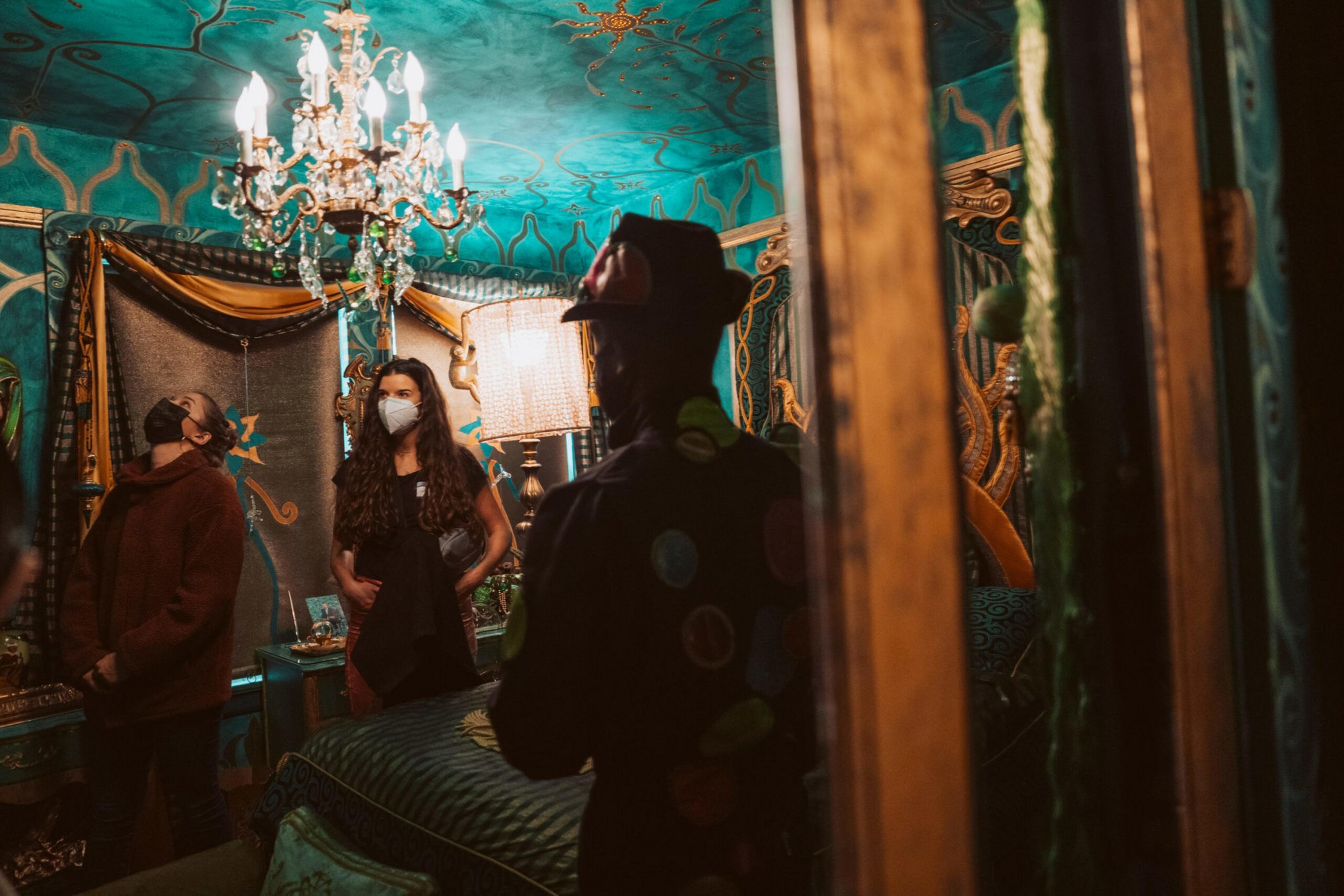 Three people wearing face masks stand in an ornately decorated room with turquoise walls, a crystal chandelier, and a large, elaborate bed.