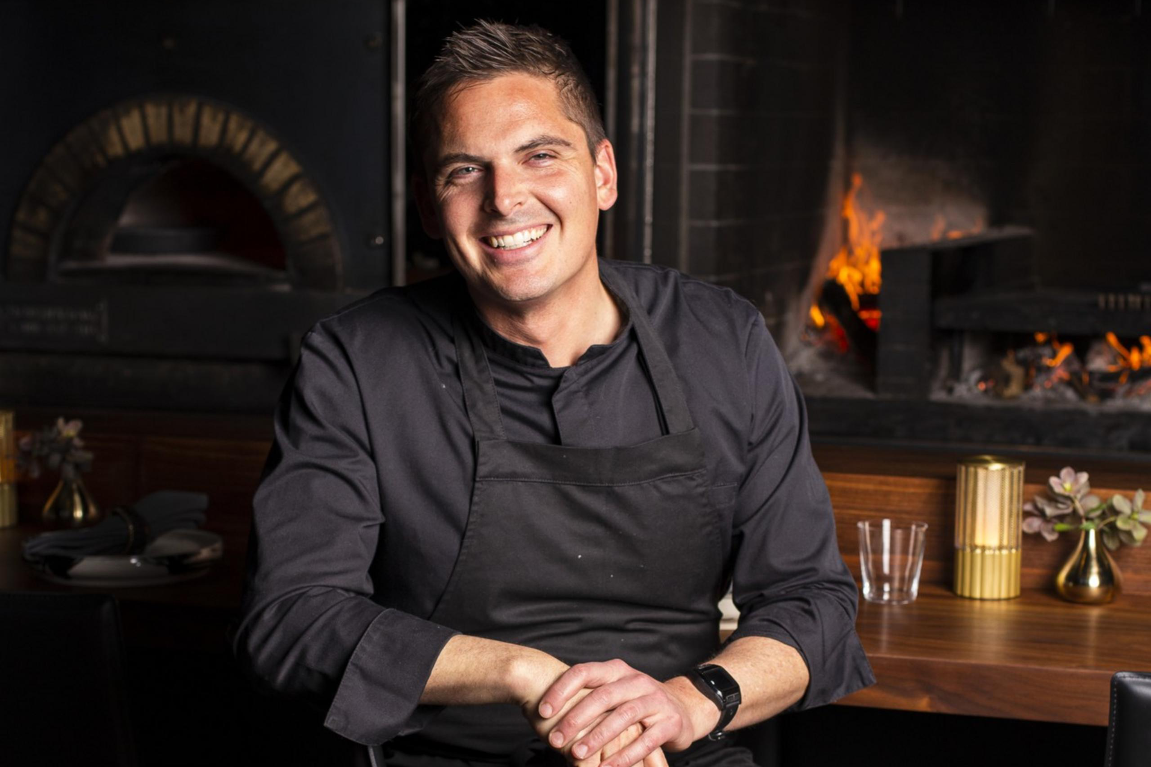 A smiling man in black chef attire sits in front of a wood-fired oven, with a fire burning. He rests his hands on his lap, creating a welcoming atmosphere.