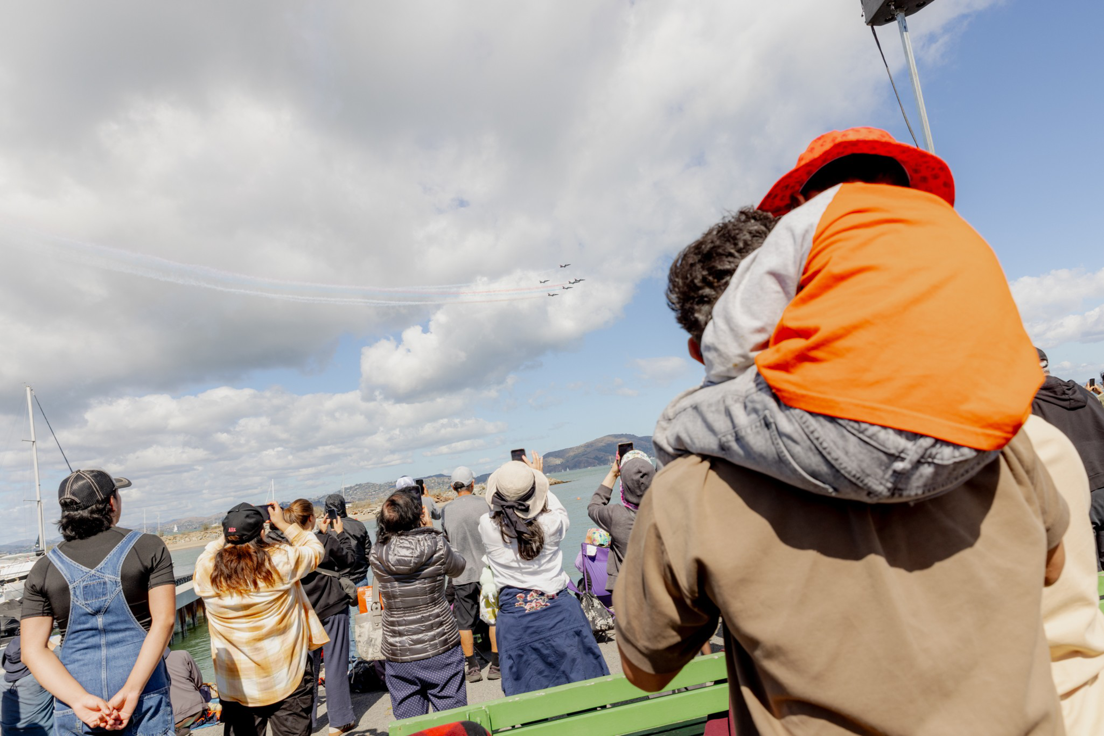 A crowd watches a blue sky air show where four planes fly in formation, leaving white trails as a child in a bright red hat sits on an adult’s shoulders.