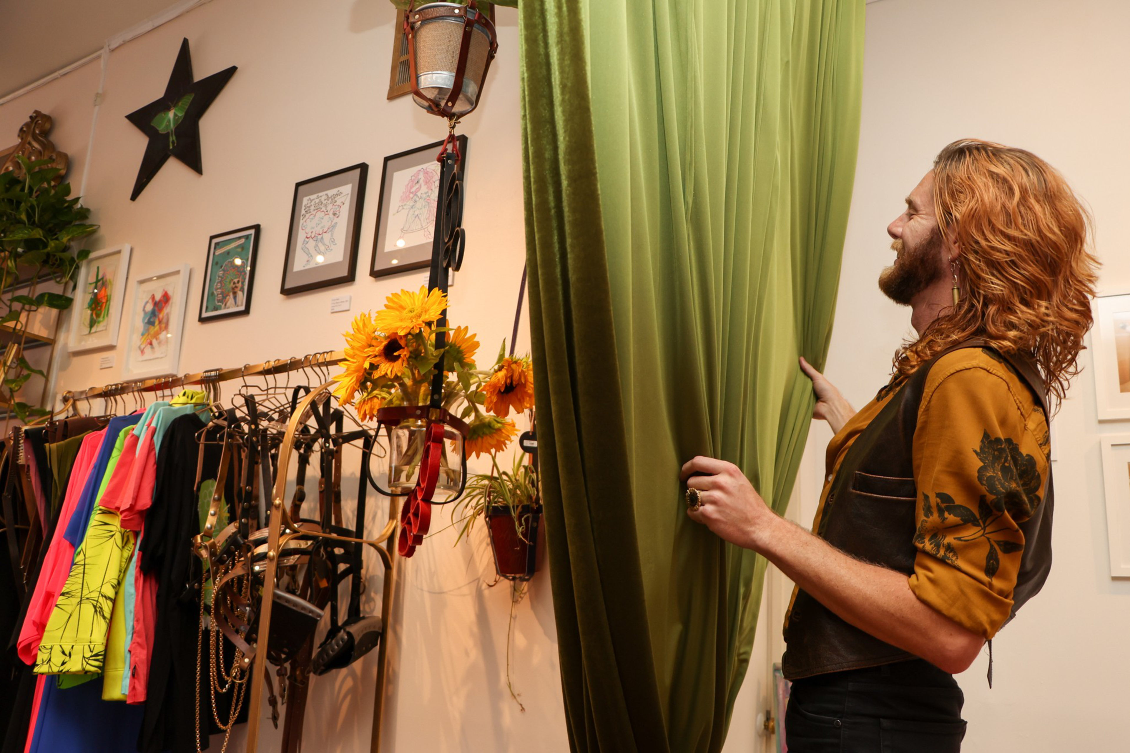 Devlin Shand, co-founder of Queer Arts Featured, hangs onto a green curtain in the art space with sunflowers and clothing in the background.