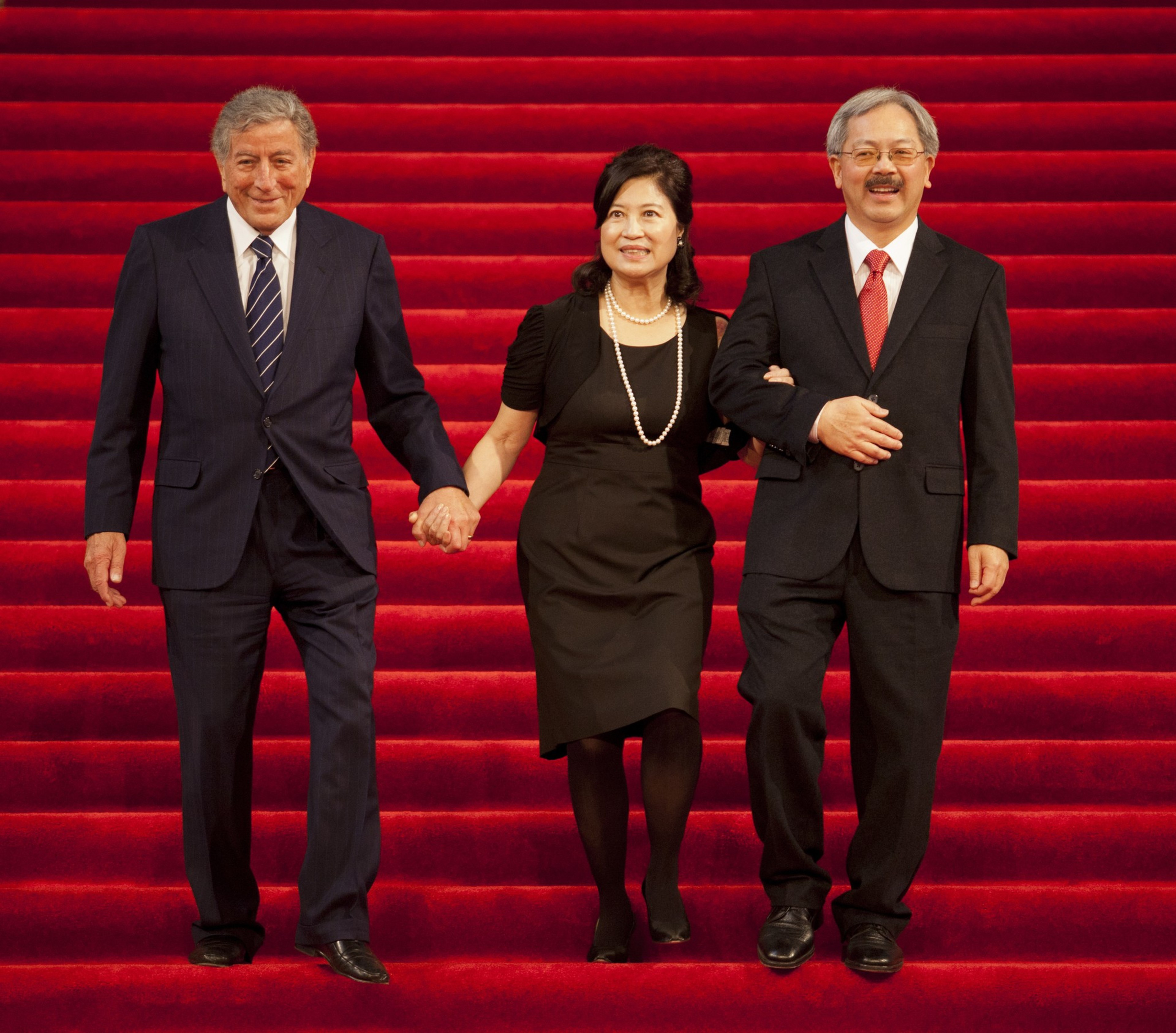 A woman in a black dress and pearls links arms with two men in suits as they walk down a wide red carpeted staircase together.