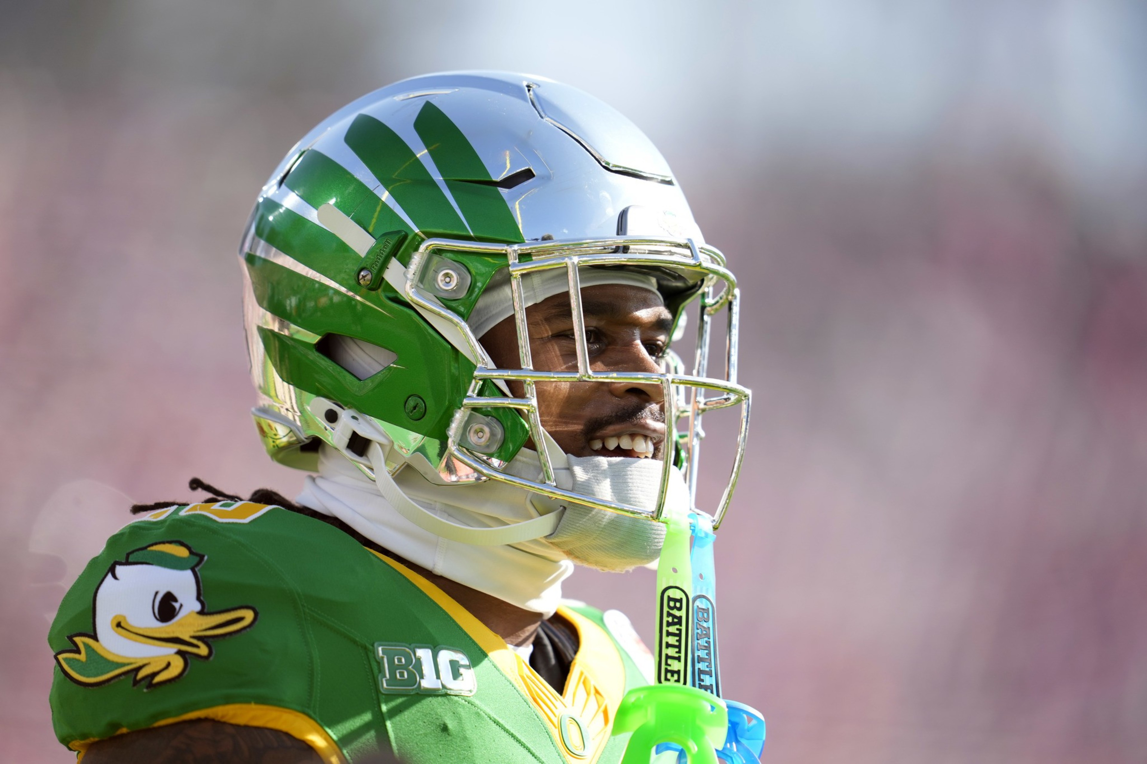 A football player in a shiny silver and green helmet smiles on the field. He's wearing a green jersey with a duck logo and holding a blue mouthguard.