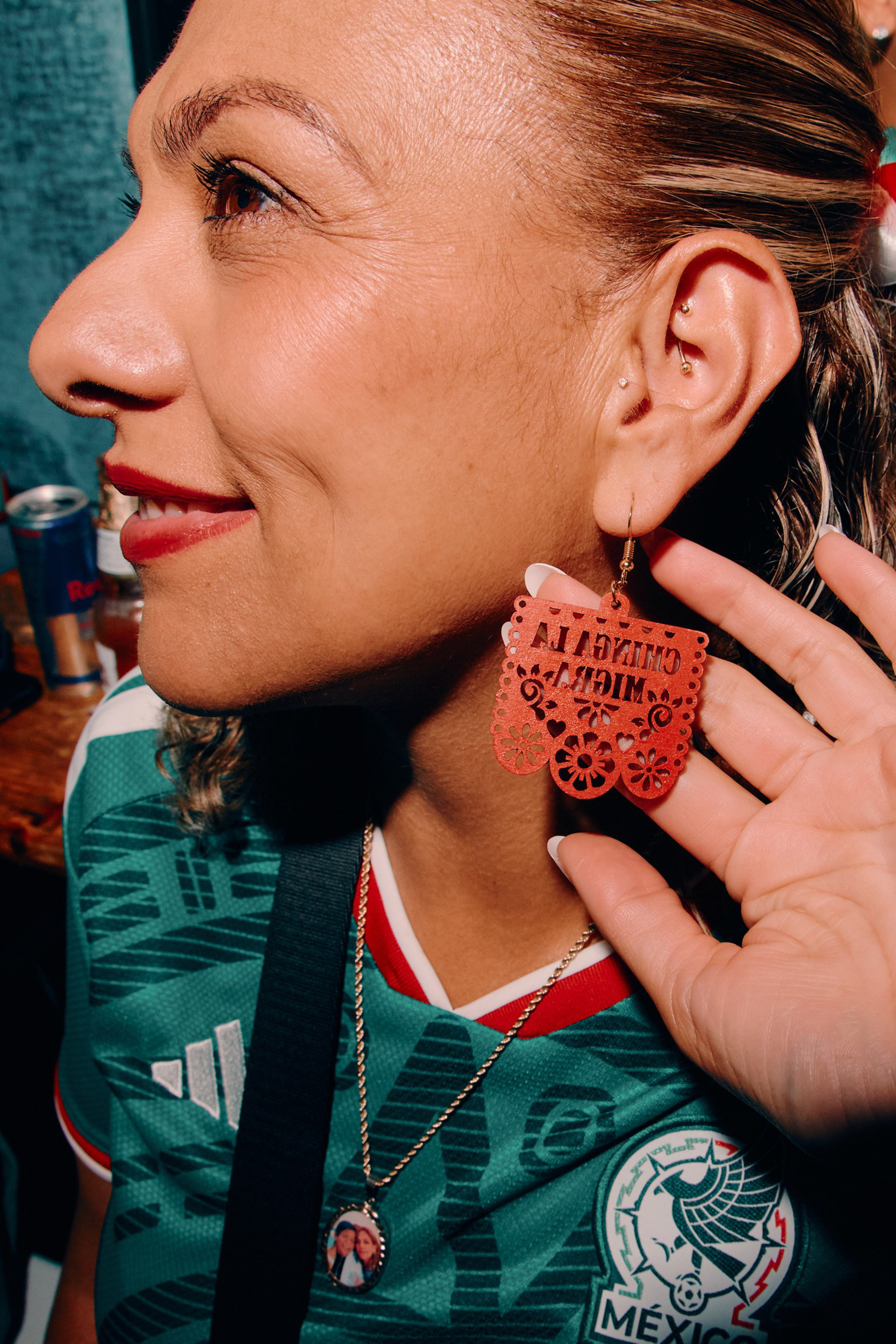 A woman wearing a green Mexico soccer jersey shows a red earring that reads “Chingona y…”. She has a necklace with a small circular photo pendant.