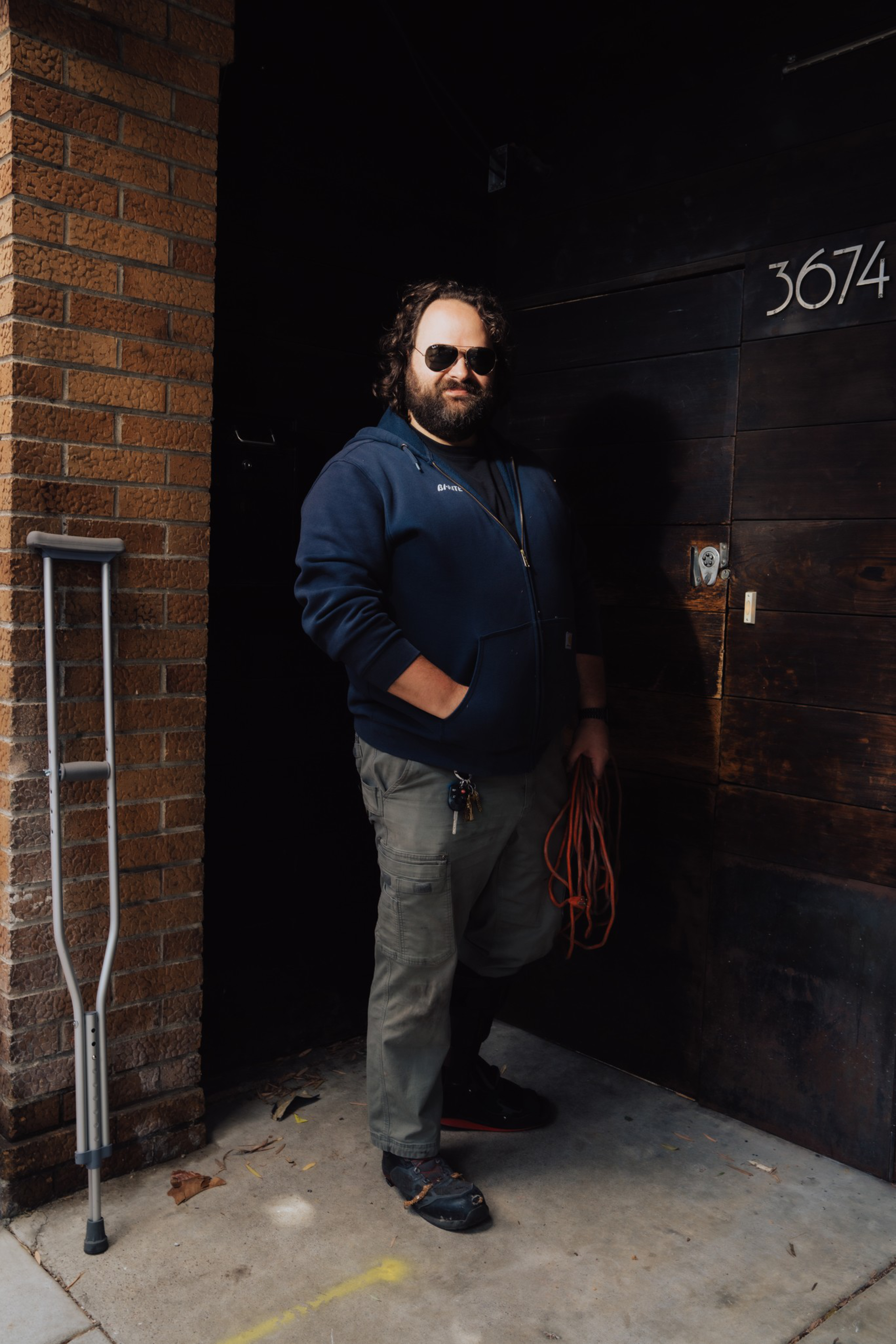 A man with a beard and sunglasses stands holding an orange extension cord, with crutches leaning against a brick wall nearby.