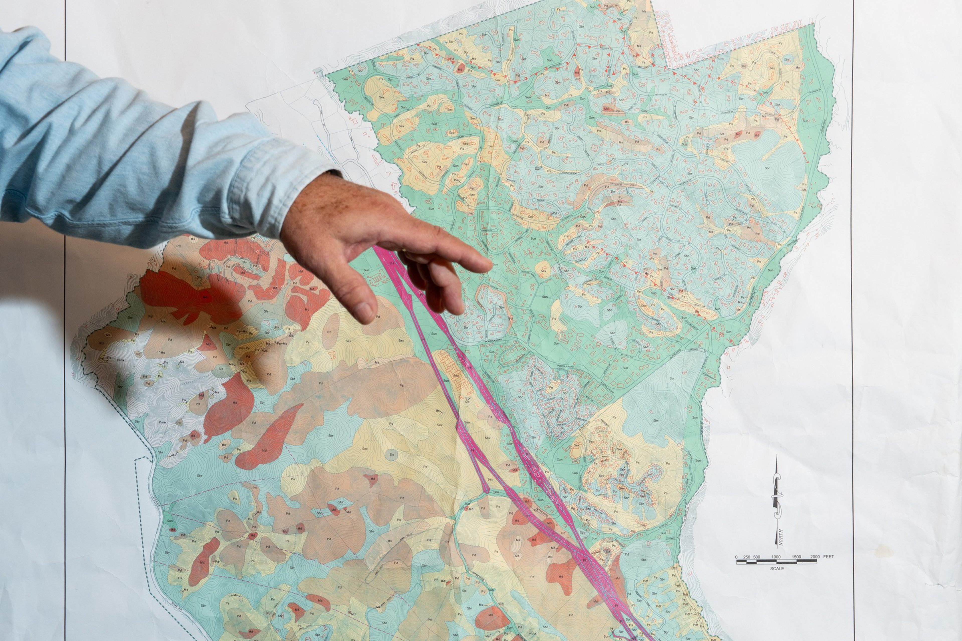 A hand points at a detailed topographic map with various colors indicating different terrains and a pink line running through the center.
